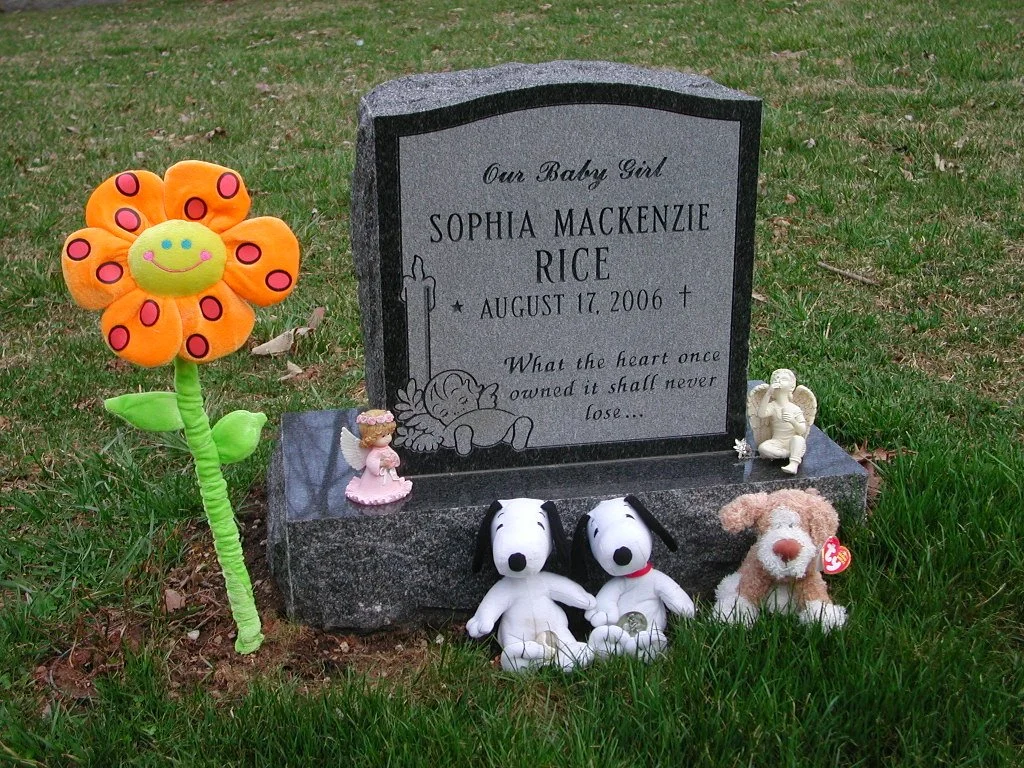 Grave marker with toys and decorations for a baby girl named Sophia Mackenzie Rice, who was born August 17, 2006, with a quote about the heart never losing. Toys include stuffed animals of Snoopy, a dog, angel figurines, and a flower-shaped plush decoration.