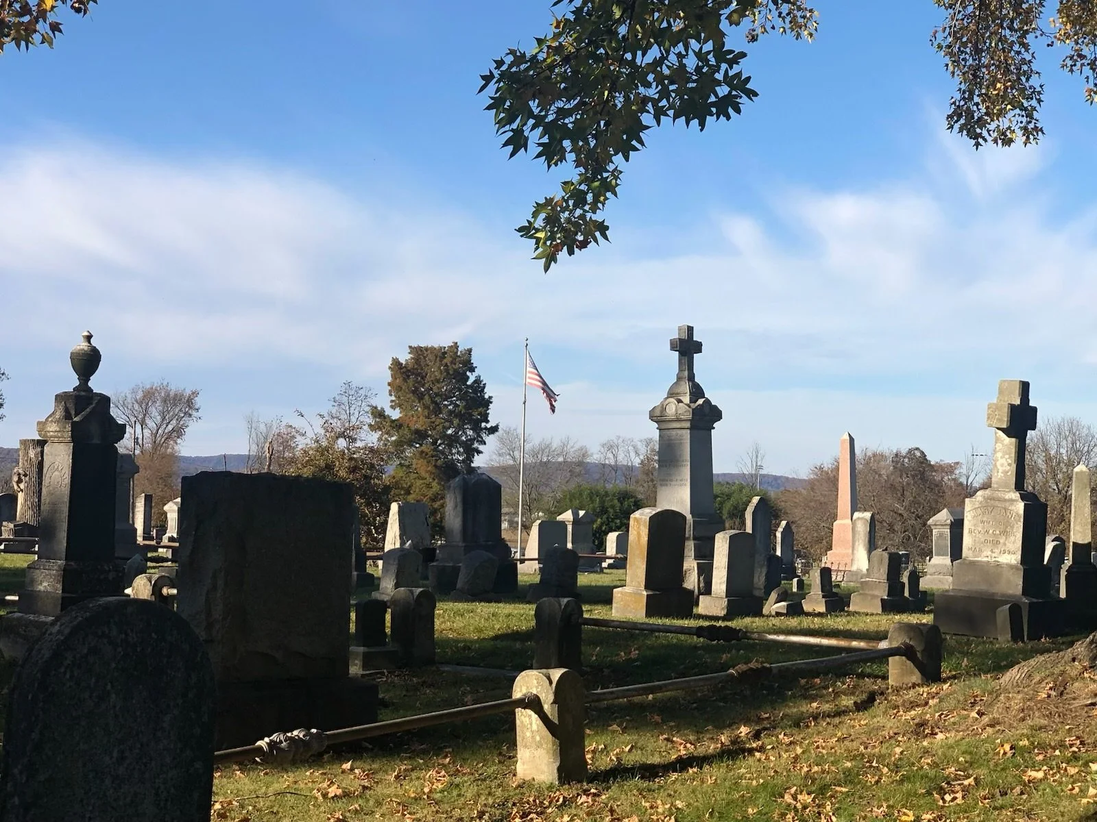 A cemetery with various tombstones and monuments, an American flag on a flagpole, and trees with autumn foliage under a blue sky with some clouds.