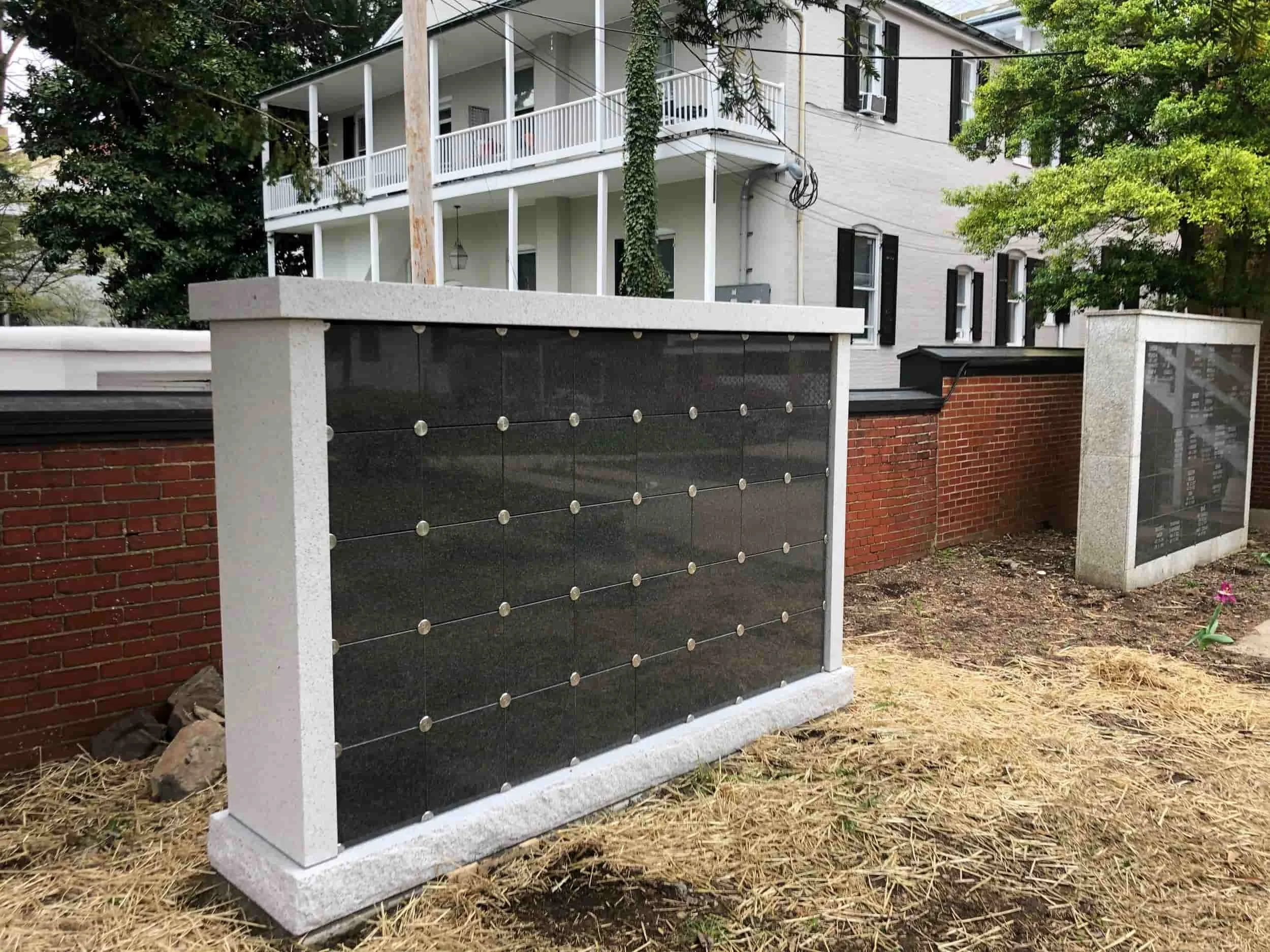 A modern black stone wall with a white concrete top, surrounded by a red brick fence with a glass panel, with a white house featuring black shutters and a balcony in the background, trees, and a chain-link fence.