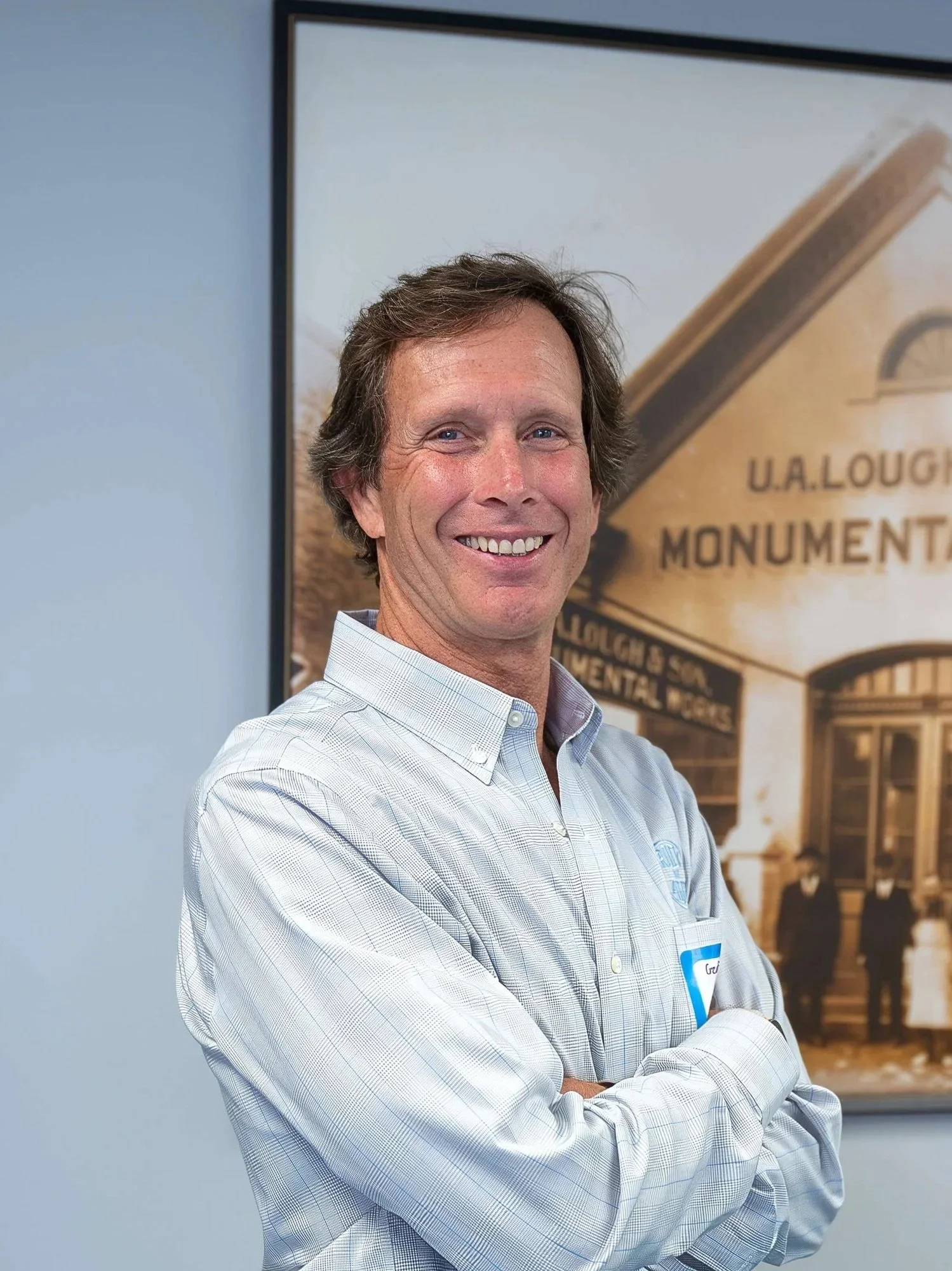 A smiling man with light skin, dark brown wavy hair, wearing a light blue checked button-up shirt, stands with crossed arms in front of a historical sign for U.A. Lougheed Monument.