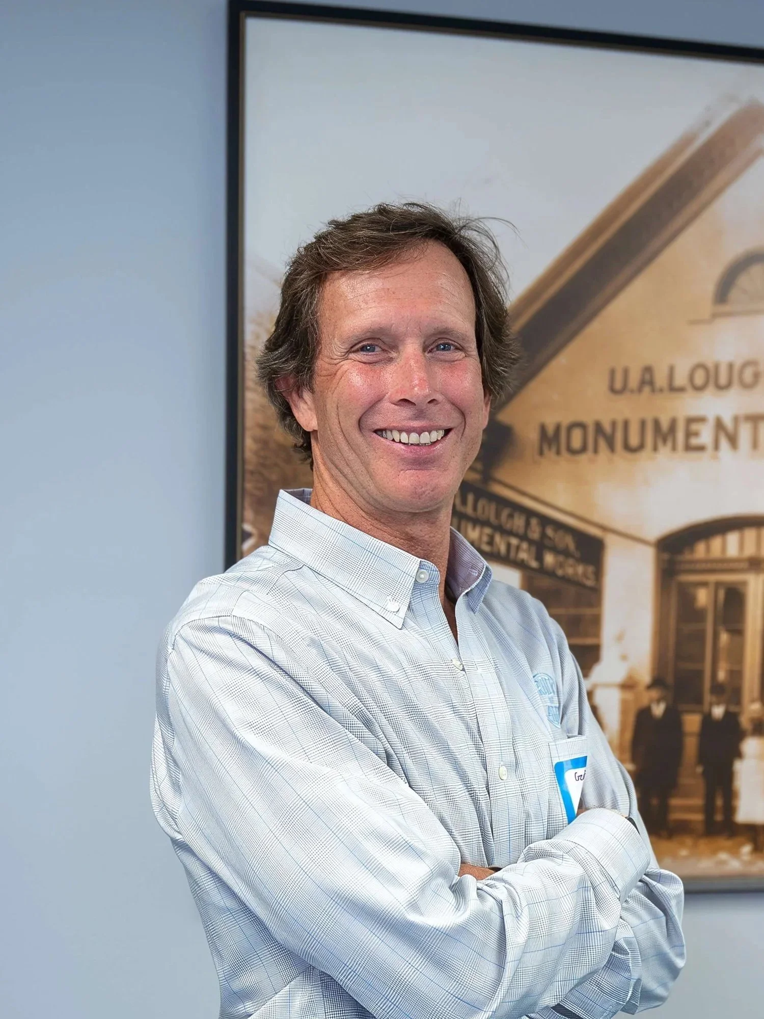 Man with light brown hair smiling, wearing a white and blue checkered shirt, standing in front of a historical photo and sign about U.A. Loug Monument and architectural details.
