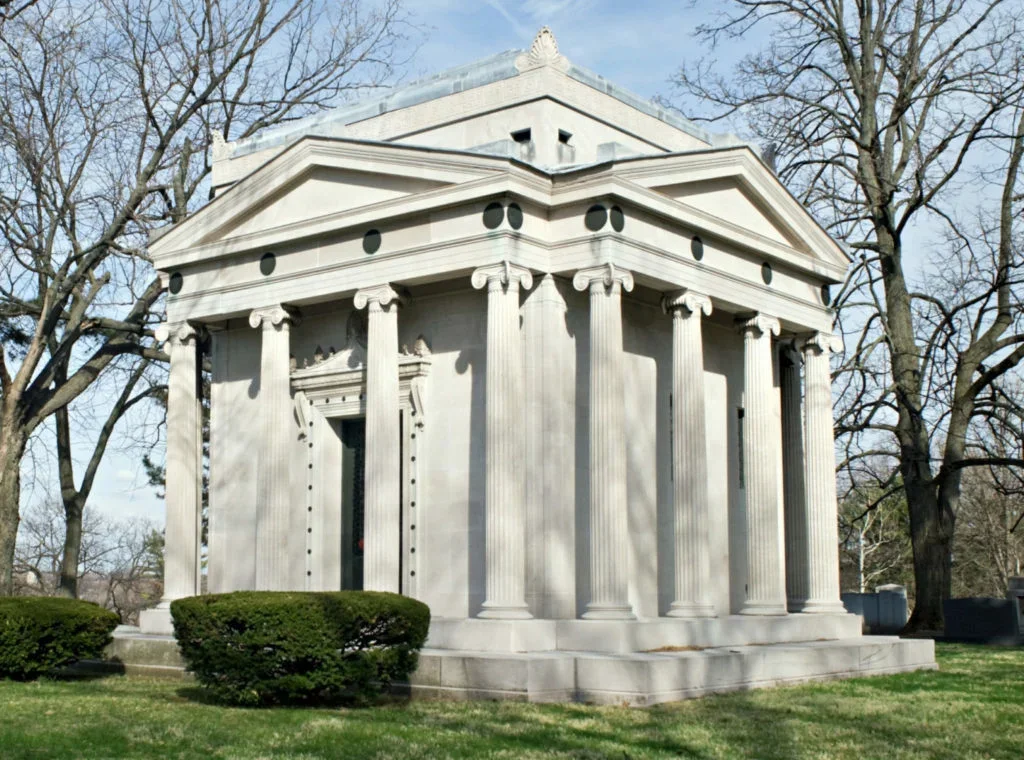 A small, white, neoclassical stone building with columns and a pediment, surrounded by grass and trees.