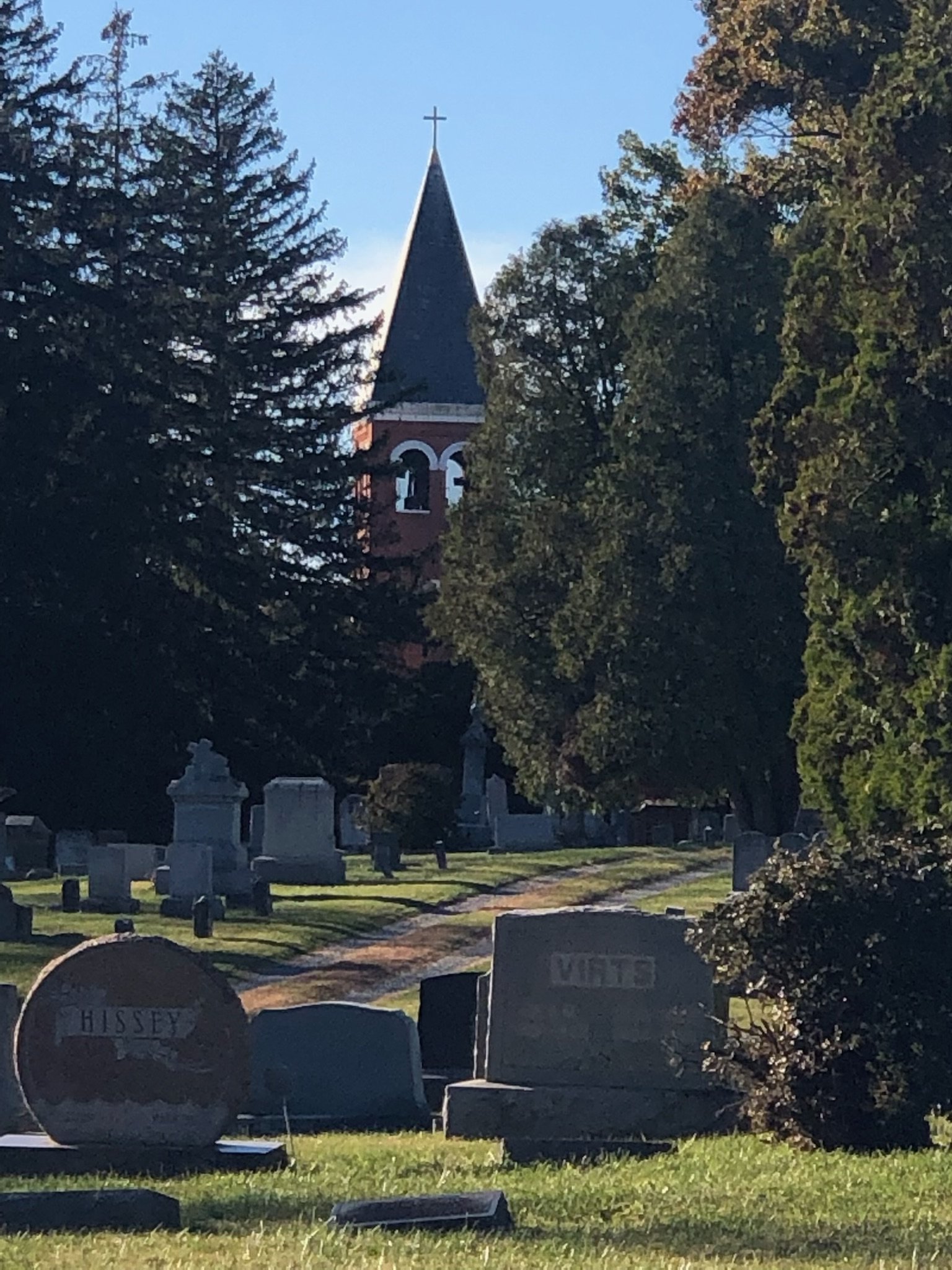 View of a cemetery with various headstones and a church steeple with a cross in the background, surrounded by green trees.