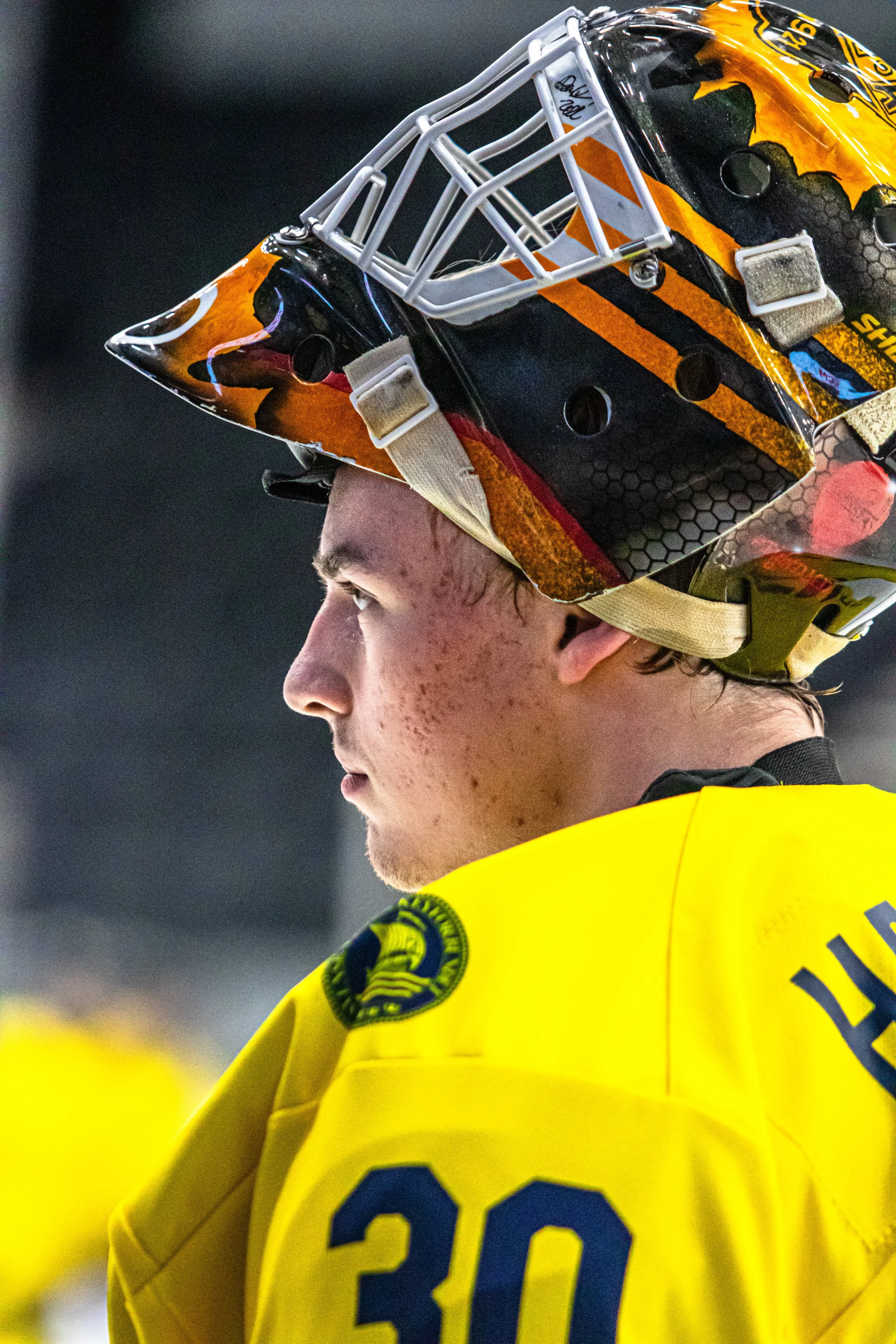 Close-up side profile of a young hockey player wearing a yellow jersey with the number 30, and a helmet with a distinctive orange and black design.