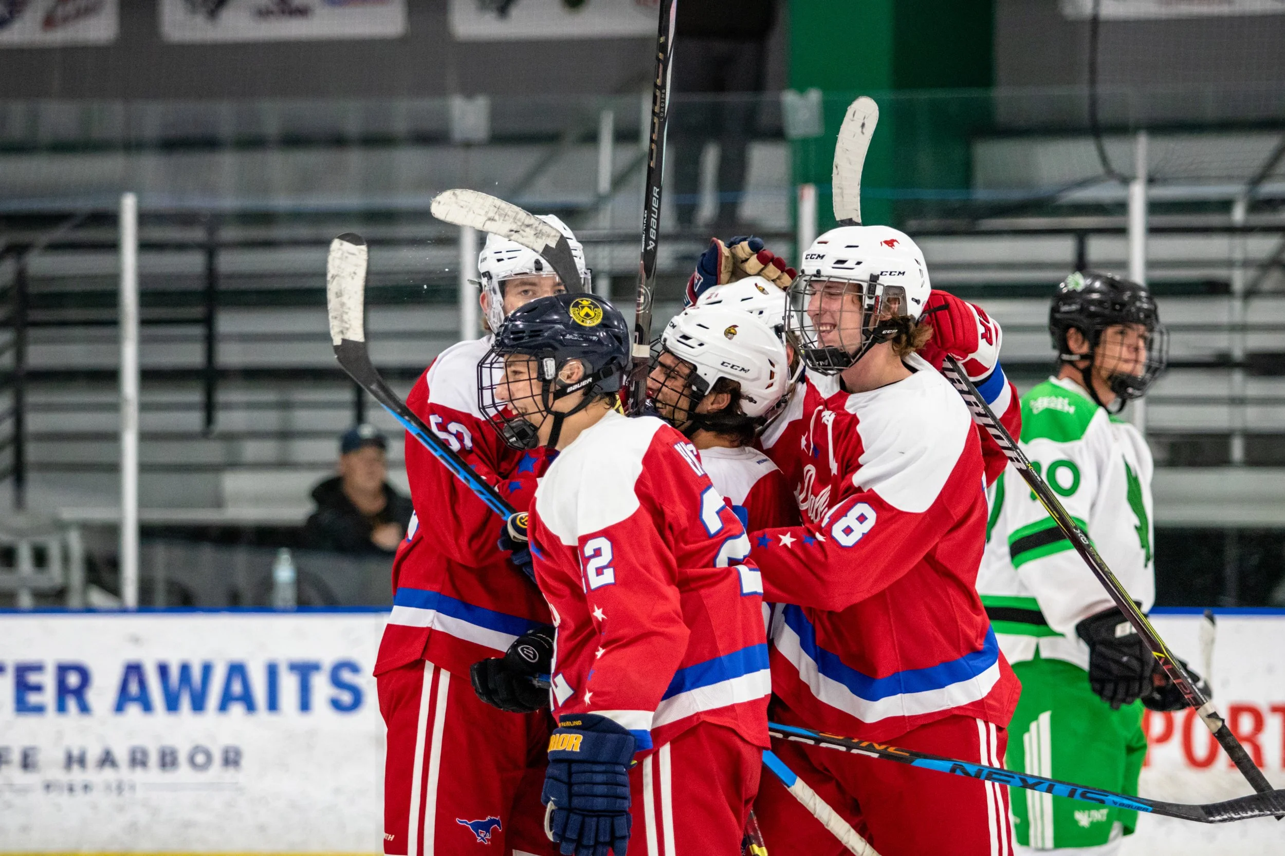 Ice hockey team in red and white jerseys celebrates together on the ice, some players are hugging and smiling, while players in green and white jerseys are in the background.