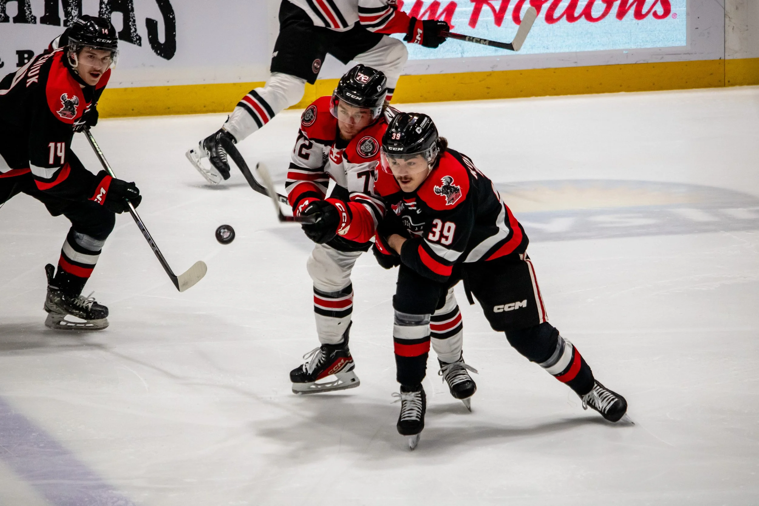 Hockey players in black and red jerseys battling for the puck on ice rink.