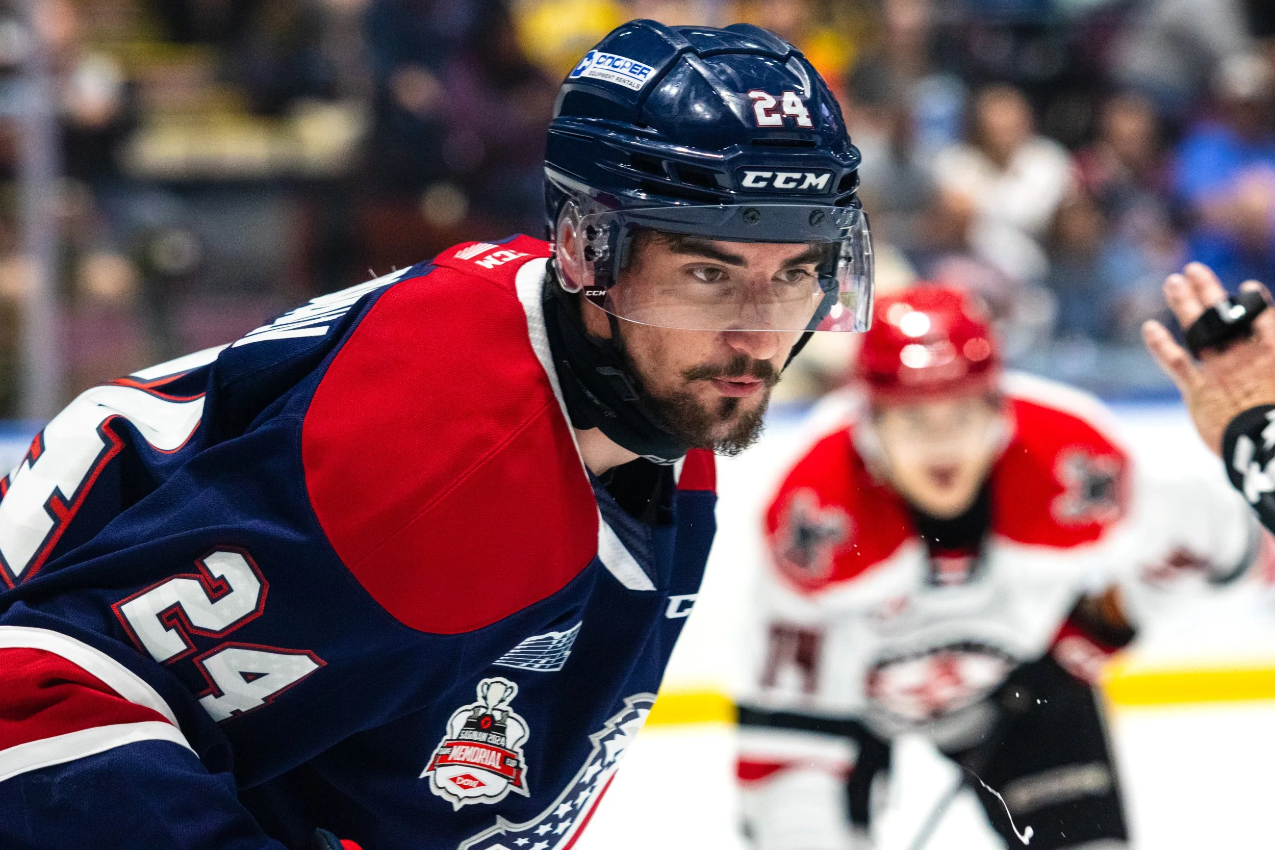 Close-up of a hockey player wearing a blue and red jersey and a dark helmet with the number 24, on the ice during a game, with another player blurred in the background.