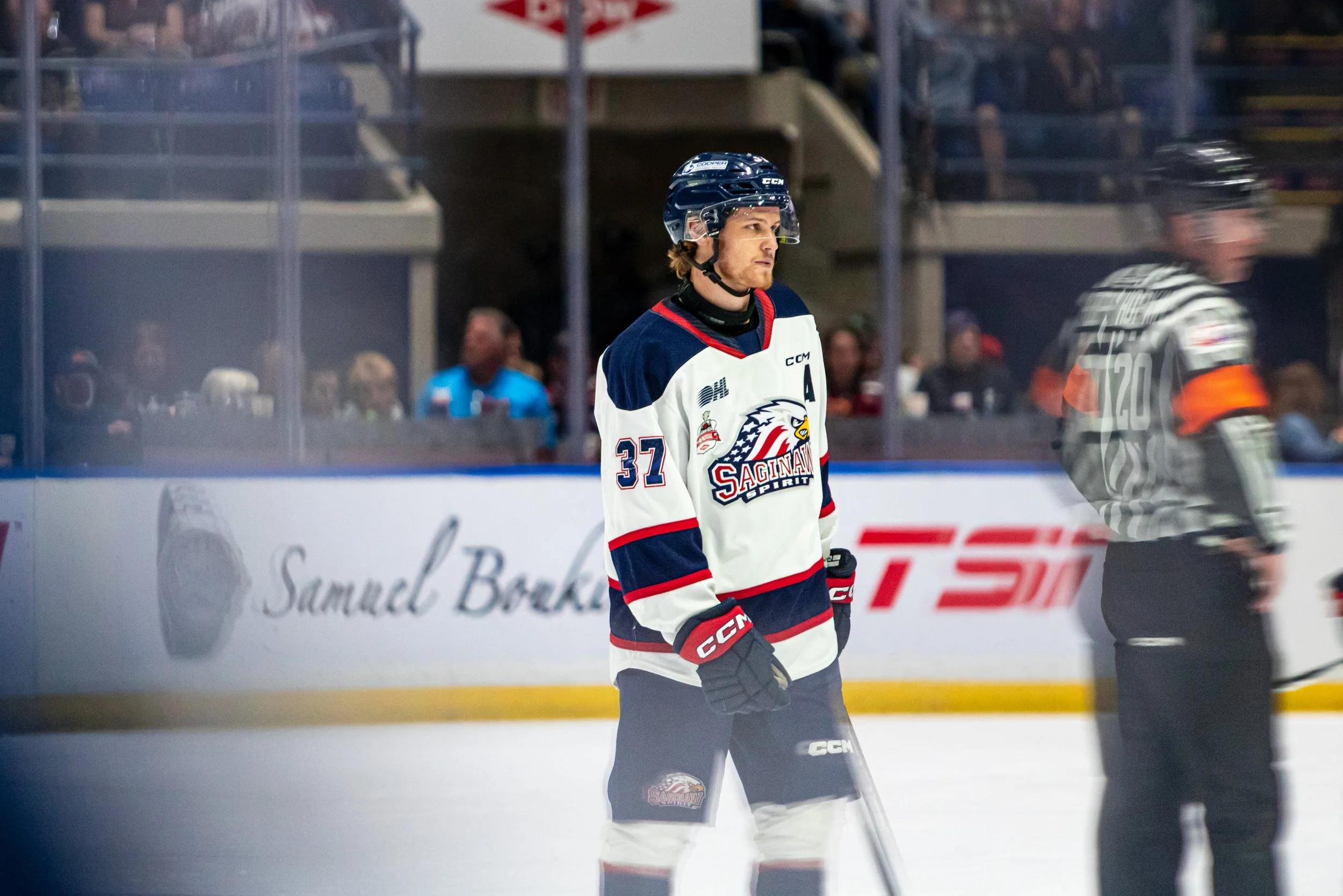 Hockey player in a Saginaw Spirit jersey standing on the ice rink with spectators in the background.