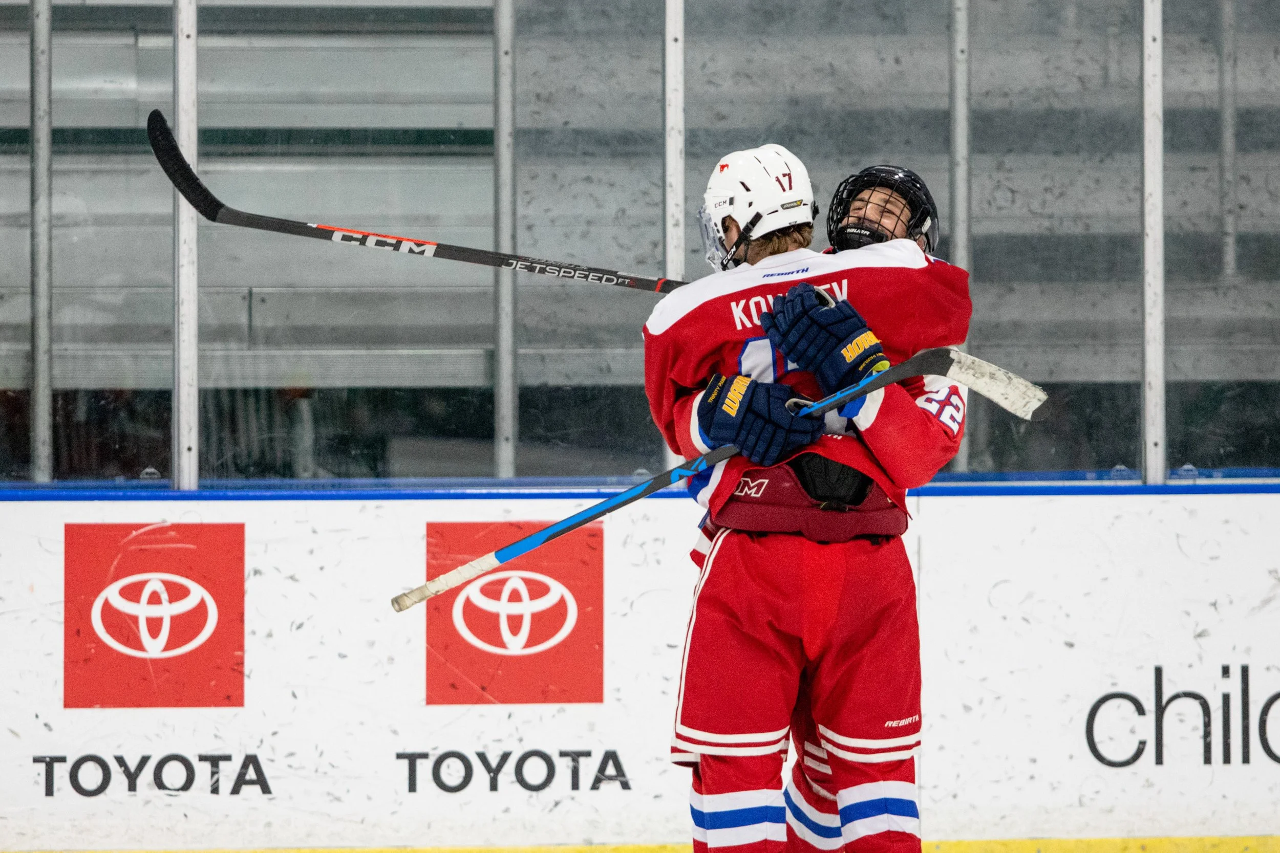 Two ice hockey players in red and white uniforms embrace after a game or play on the ice rink.