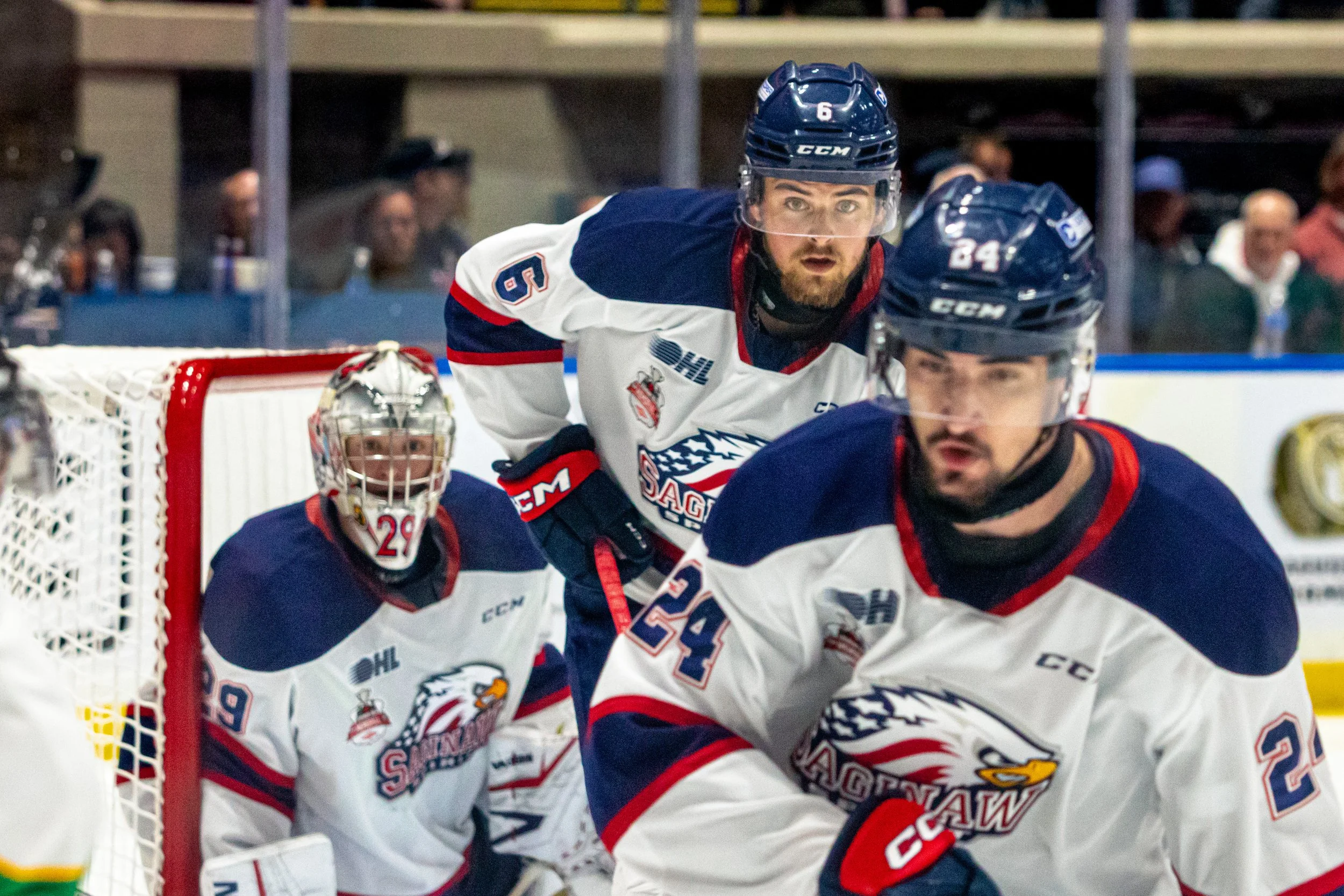 Ice hockey players in white and navy jerseys on the ice during a game, with a goalie in the background and spectators watching in the stands.