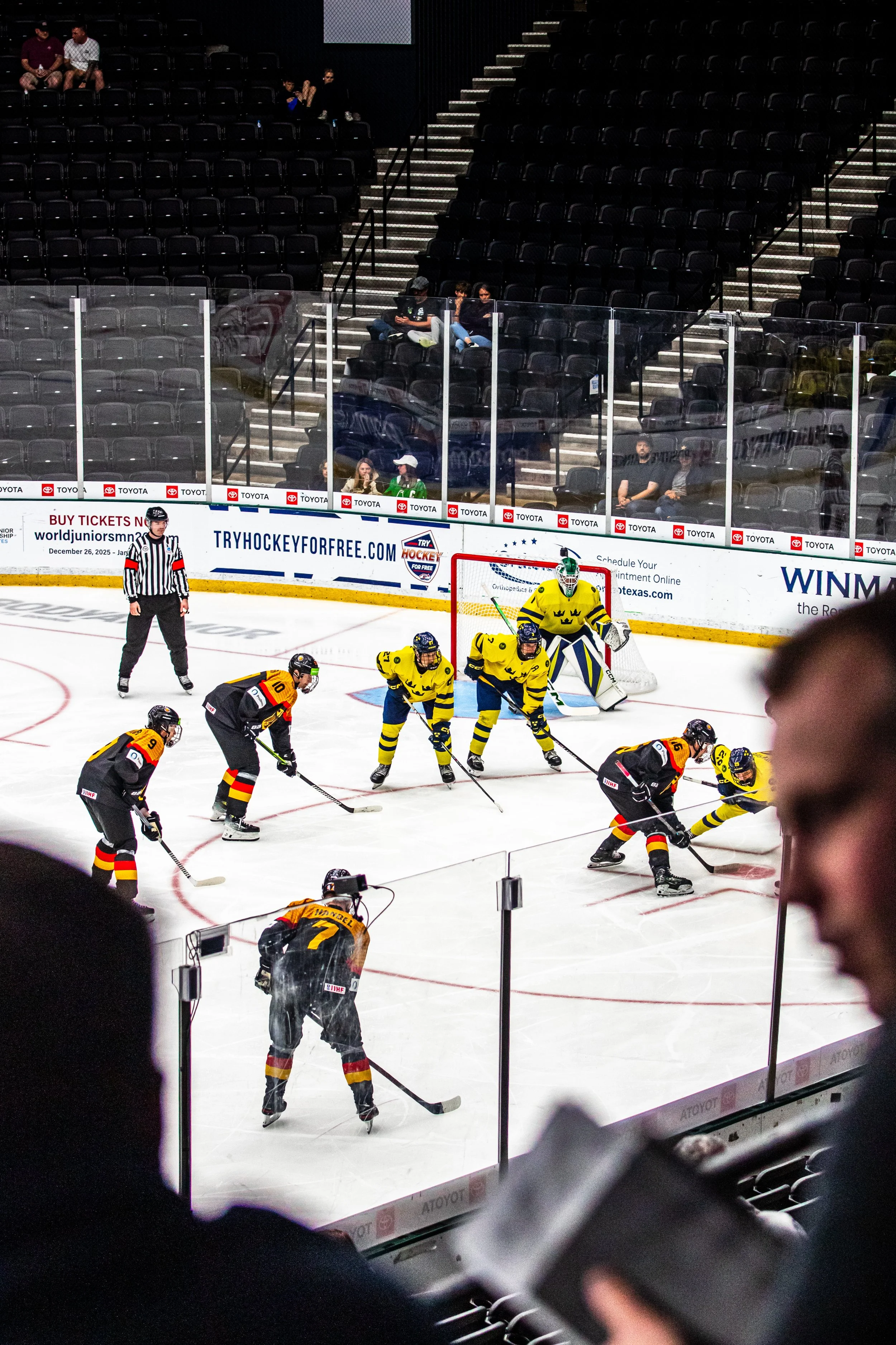 Ice hockey game in progress with players from two teams, one in yellow and blue jerseys and the other in black and red jerseys, near the goal. Spectators sit in the stands behind protective glass.