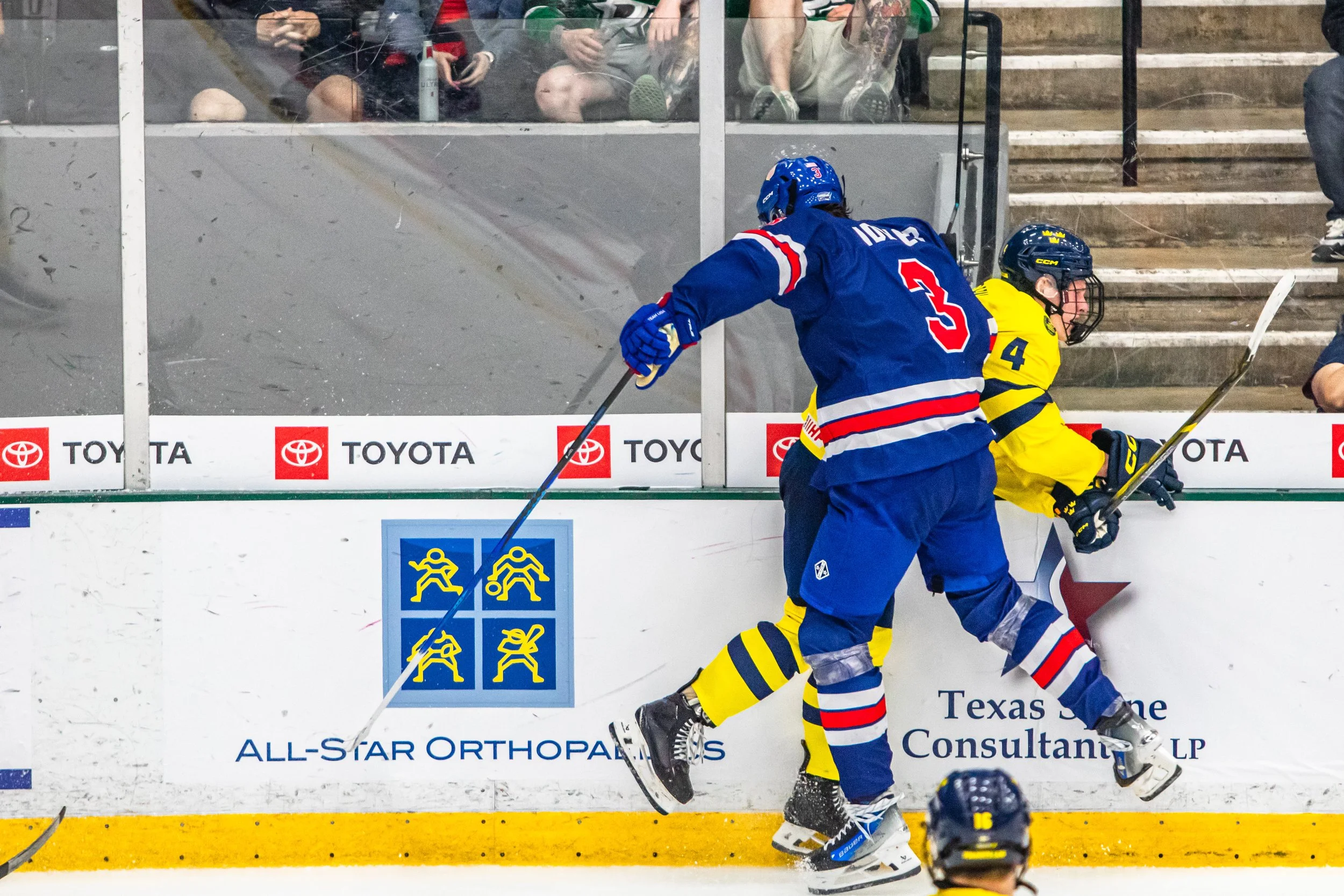 Hockey players in a fight along the rink boards, one in a blue uniform and the other in a yellow uniform, with spectators watching from the stands.