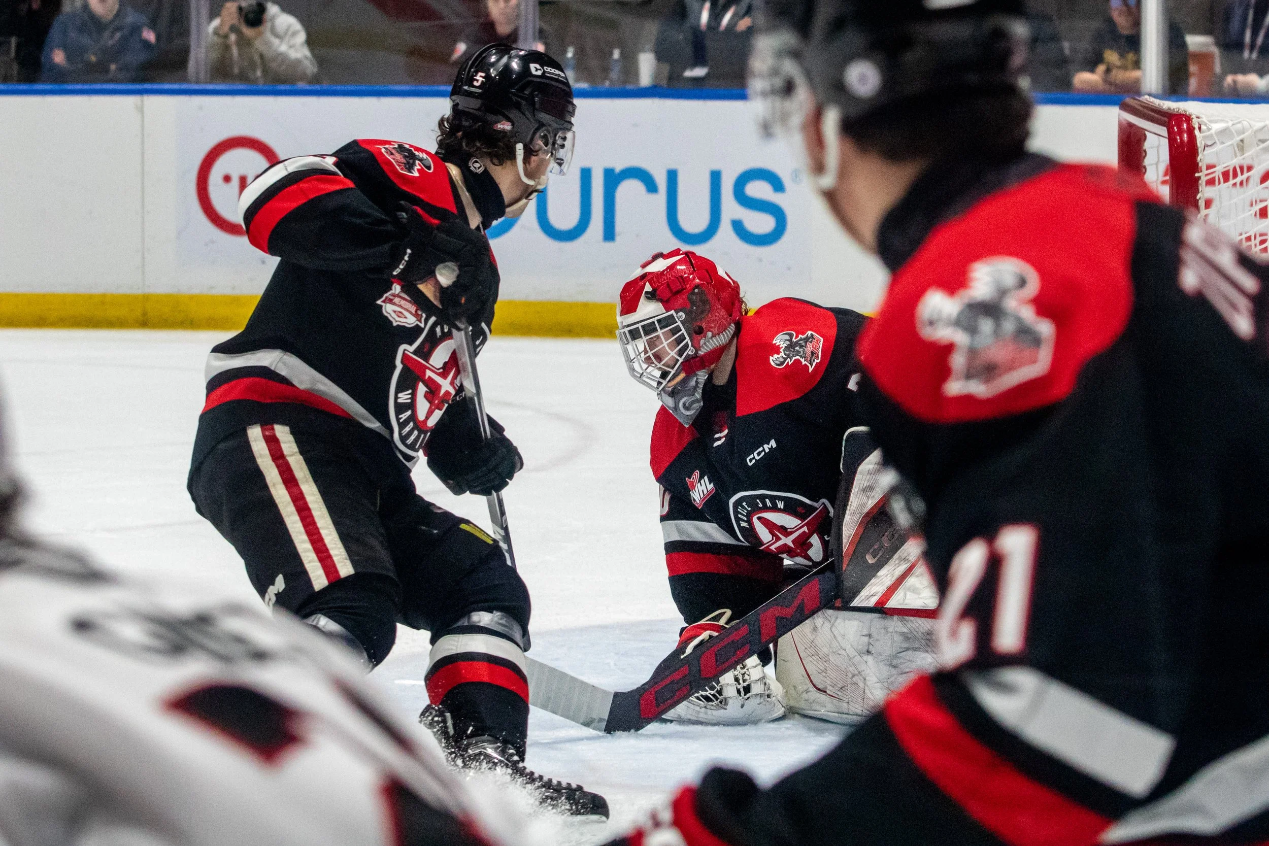 Hockey players in black and red uniforms on ice, one player appears to be a goalie in goalie gear, with other players surrounding him near the goal.