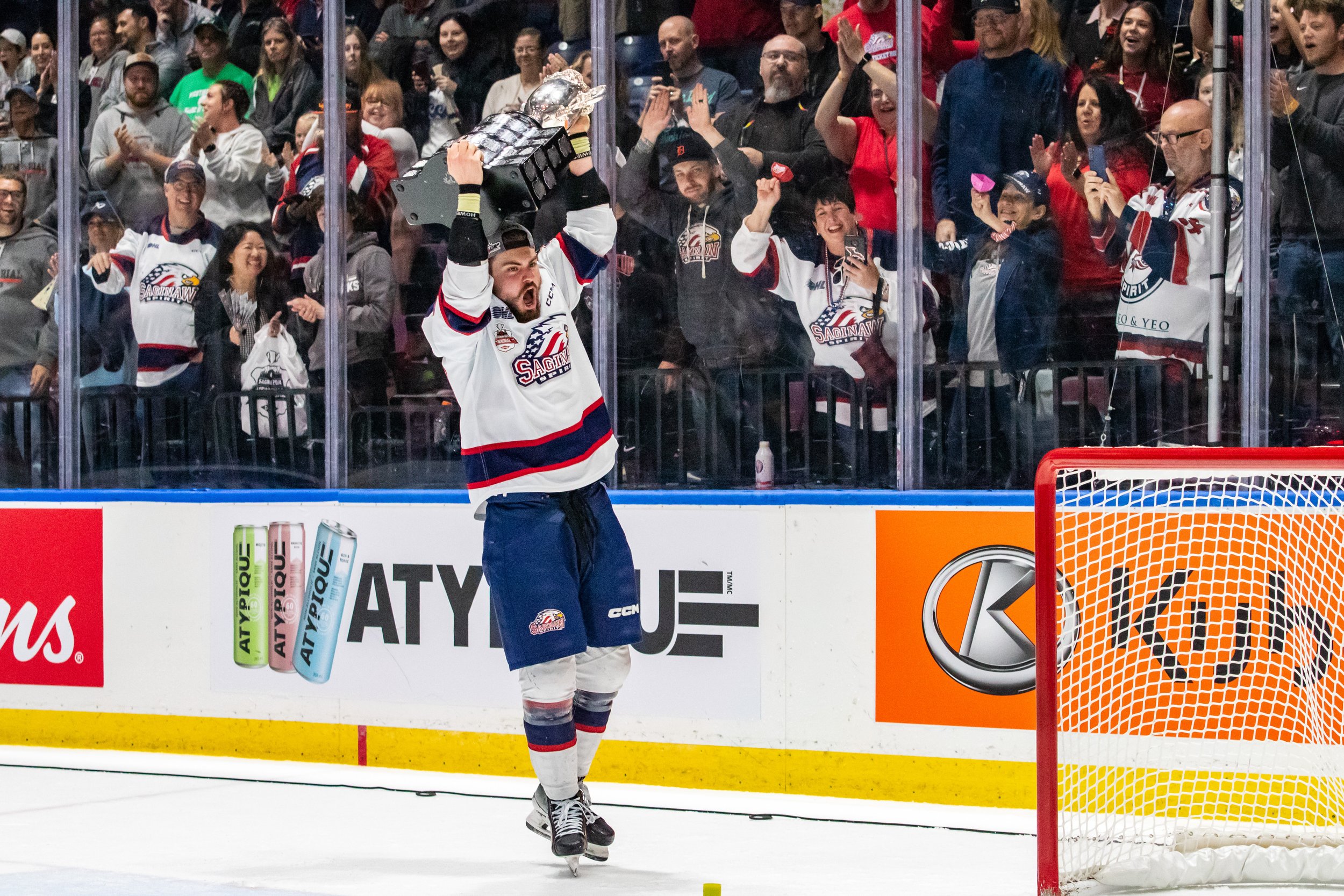 Hockey player in a white jersey with blue and red stripes holding a trophy above his head on the ice rink, celebrating after a game, with cheering fans behind glass boards.