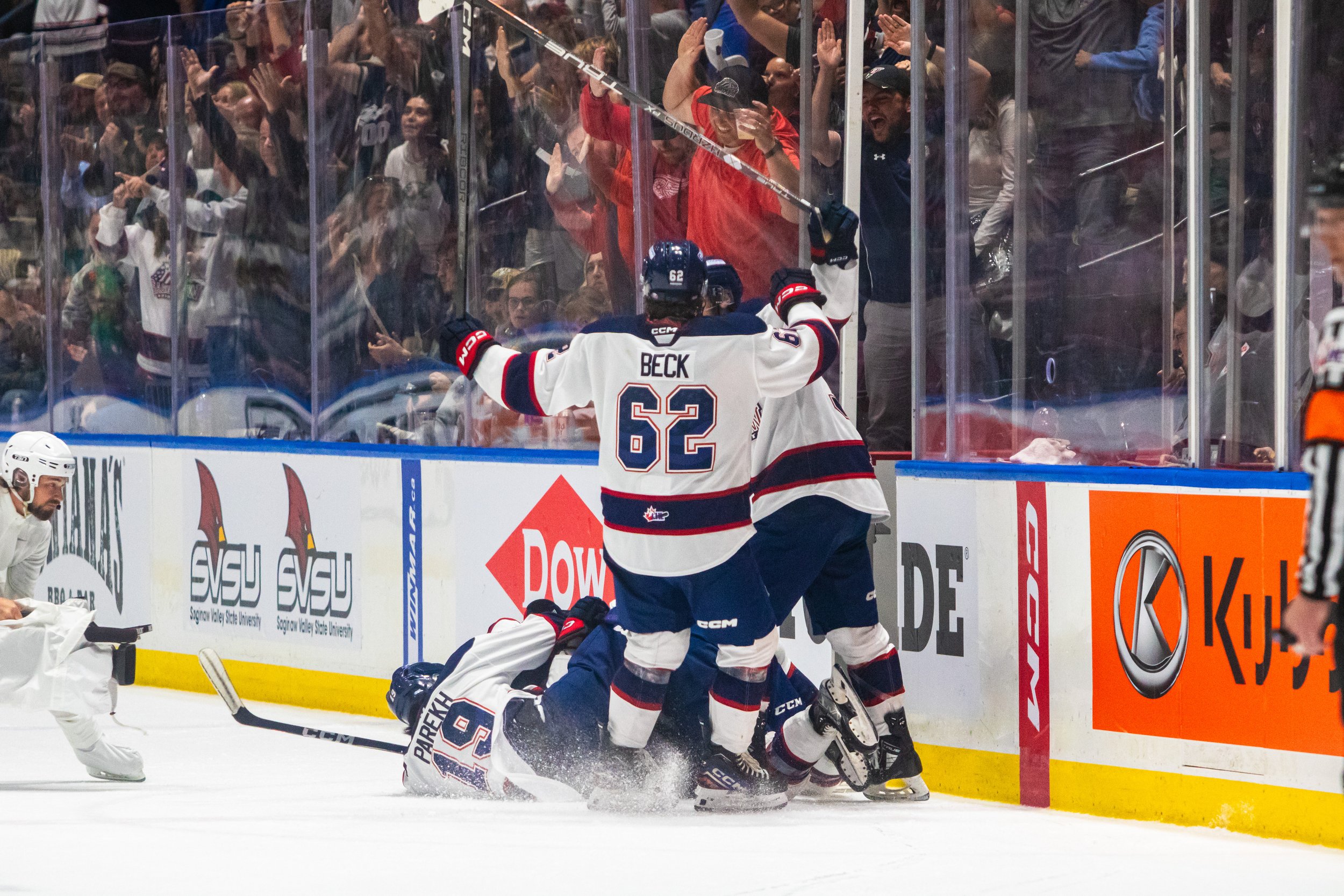 Hockey players celebrating a goal on the ice, with one laying on the ice and others jumping on top, while fans cheer behind the glass.