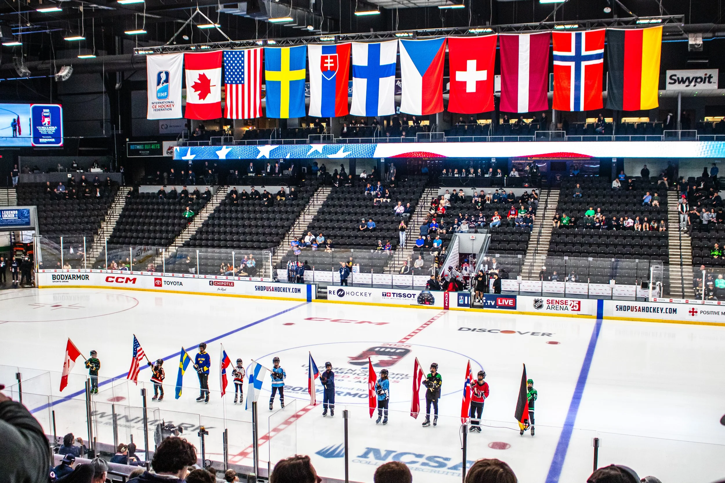 An indoor hockey arena with flags of various countries hanging above the ice rink. Players are lined up on the ice holding flags representing different nations, with spectators seated in the stands watching the event.