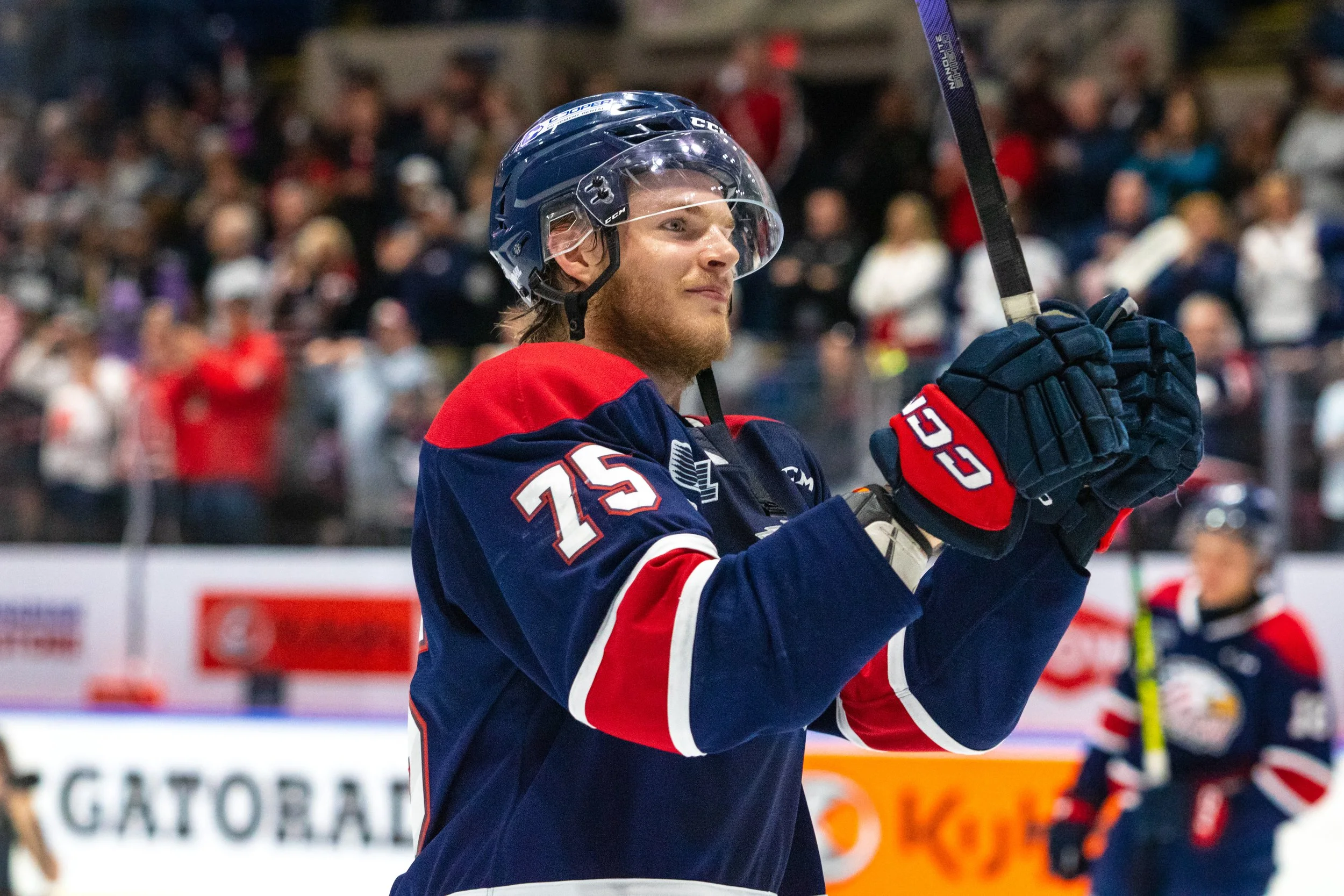 A hockey player wearing a blue and red jersey with the number 75, holding a hockey stick, standing on ice rink.