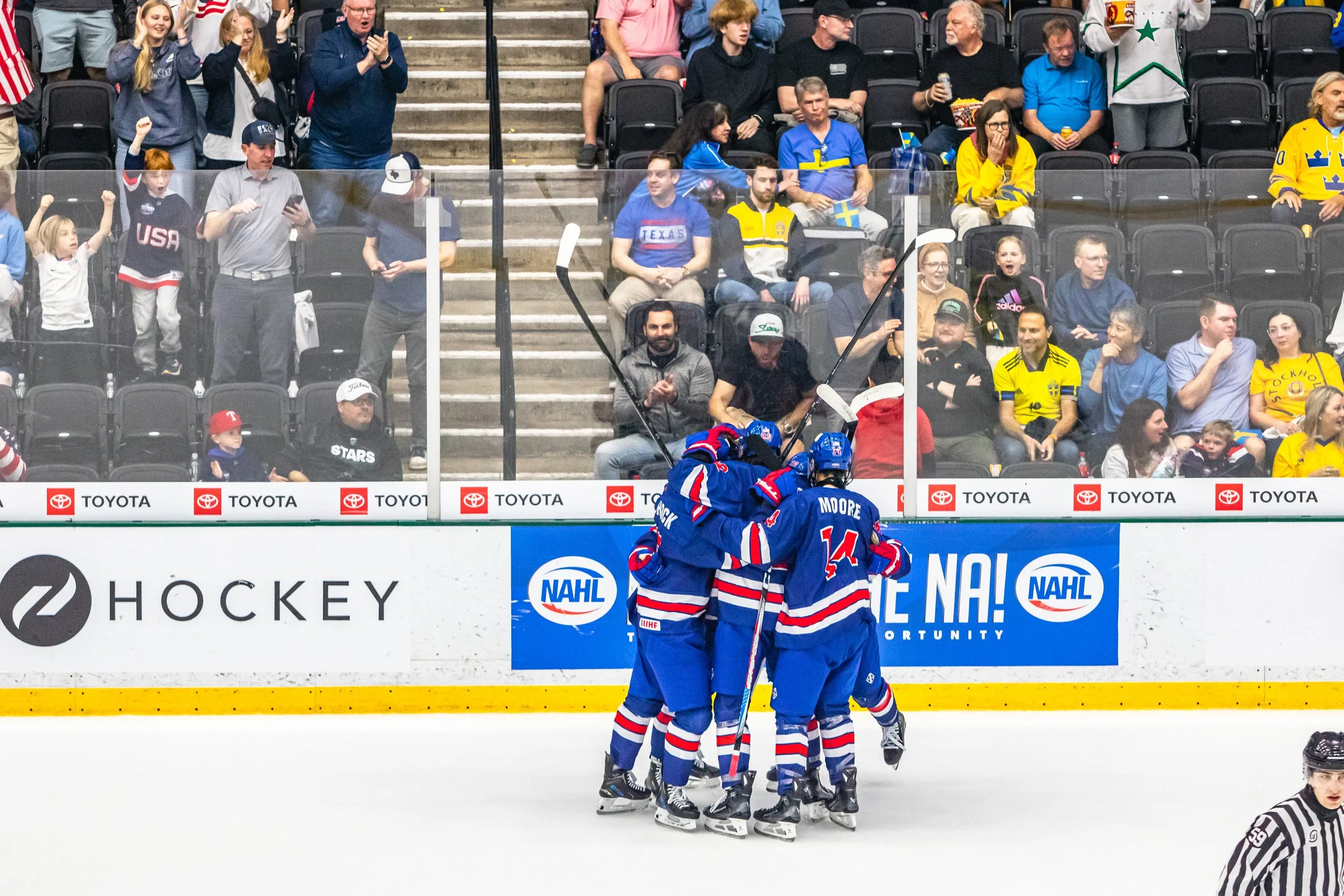 Hockey players in blue jerseys celebrate a goal on the ice rink while fans cheer in the stands behind the glass, with some fans wearing Swedish and American team apparel.