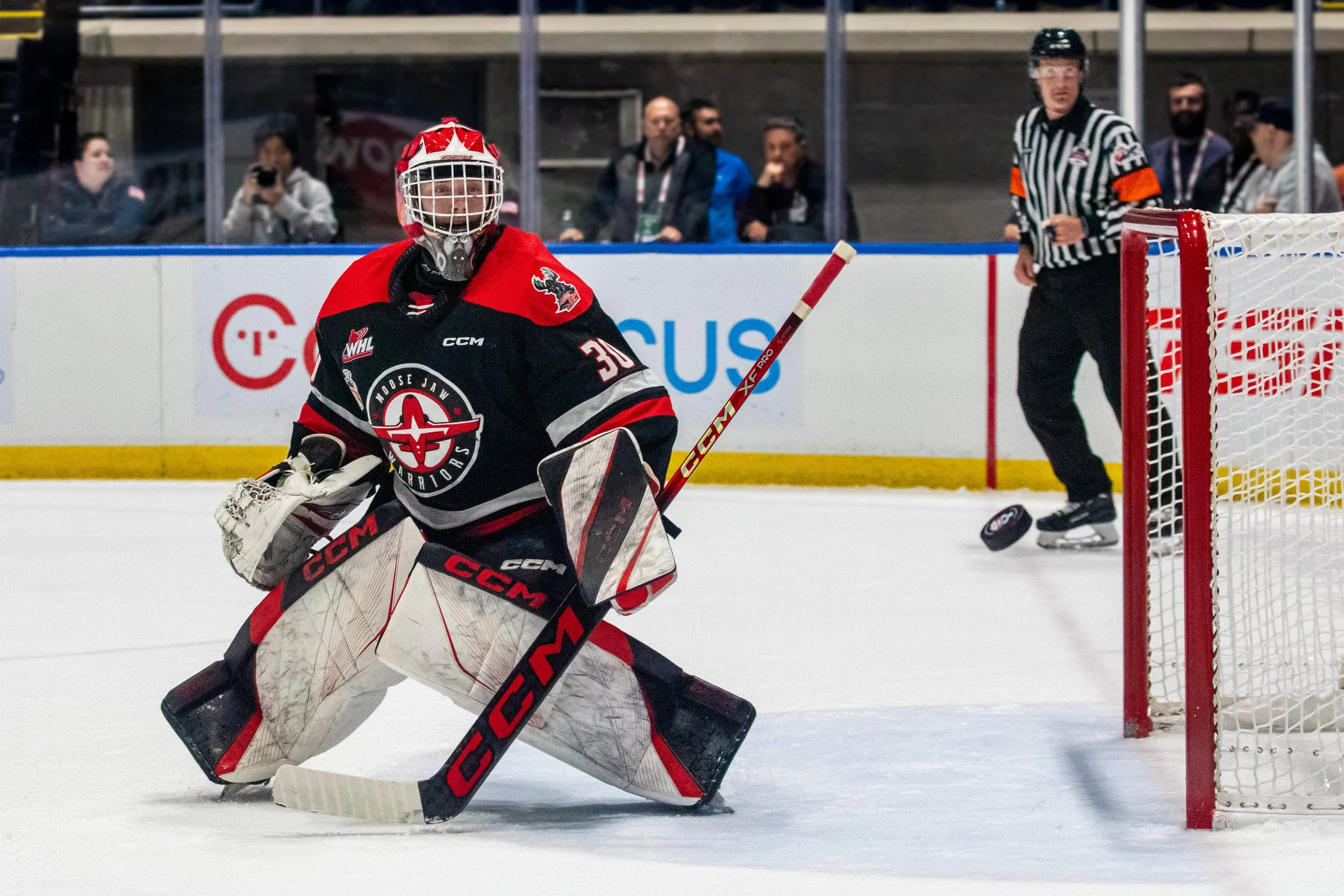 An ice hockey goalie in black and red uniform preventing a puck from entering the net during a game.