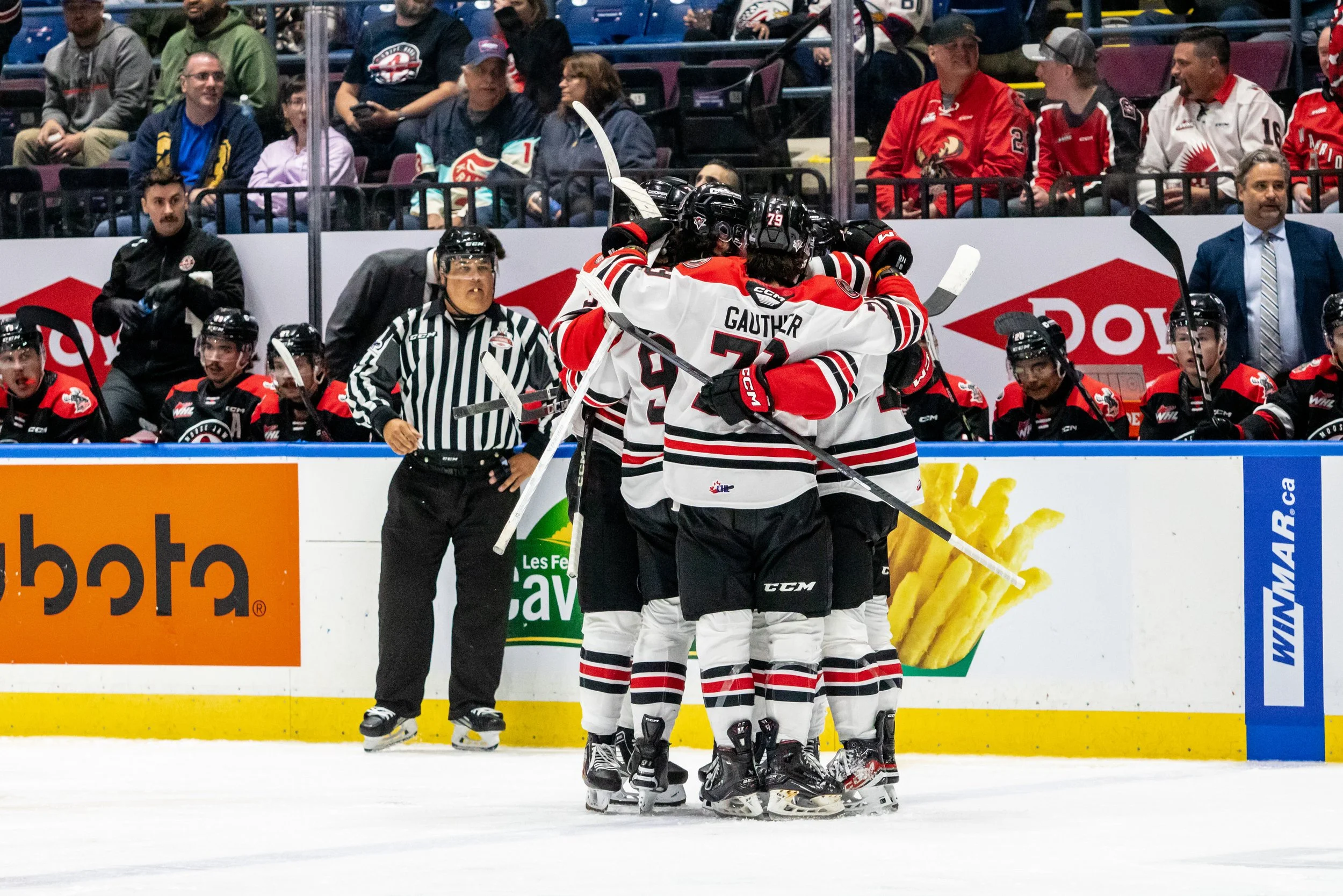 Hockey players in white, red, and black jerseys celebrate by hugging on the ice during a game. Spectators watch from the stands.