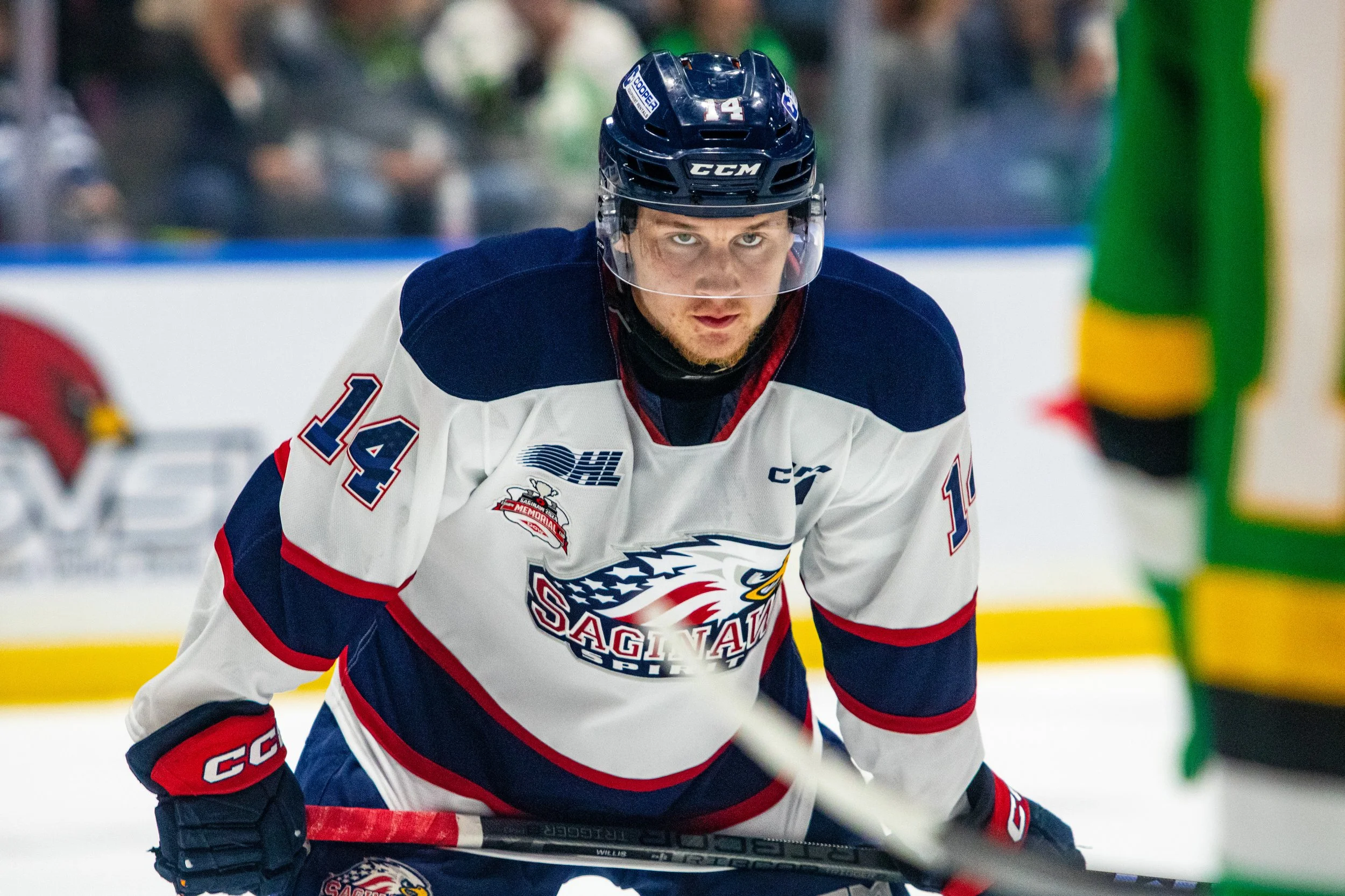 A male ice hockey player wearing a white and blue jersey with the number 14, crouched with a focused expression on his face during a game.