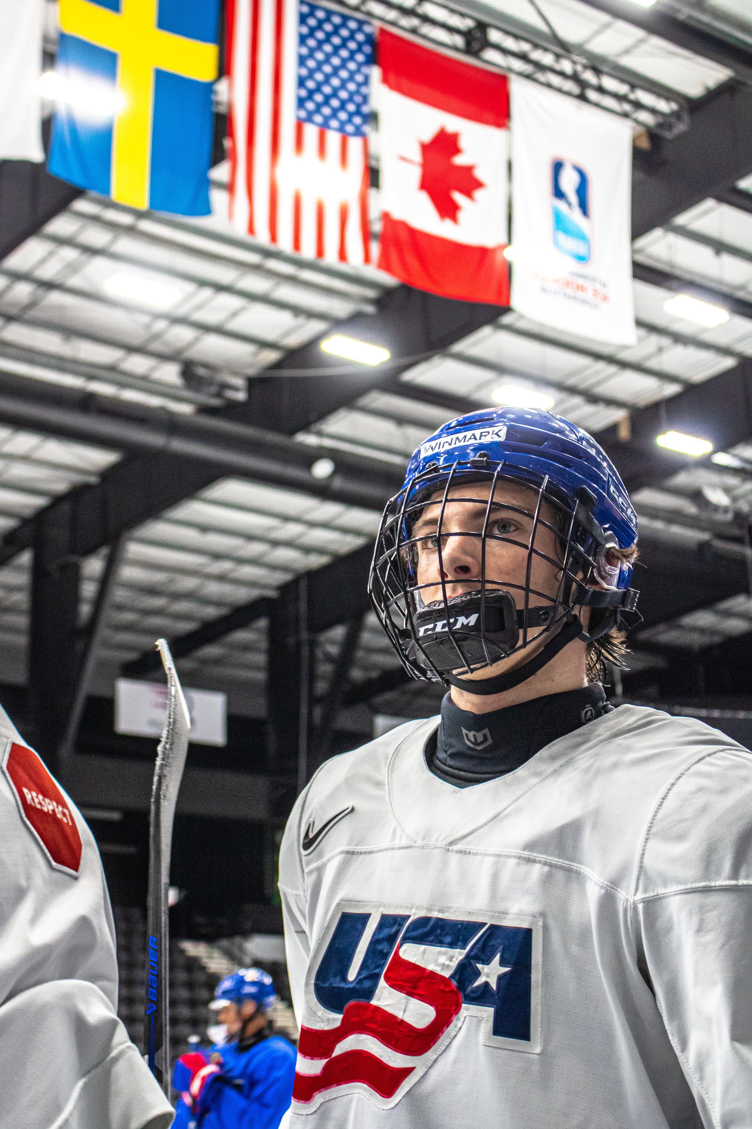 An ice hockey player wearing a USA jersey and helmet, standing inside an ice rink with flags of the USA, Sweden, Finland, and Norway hanging in the background.