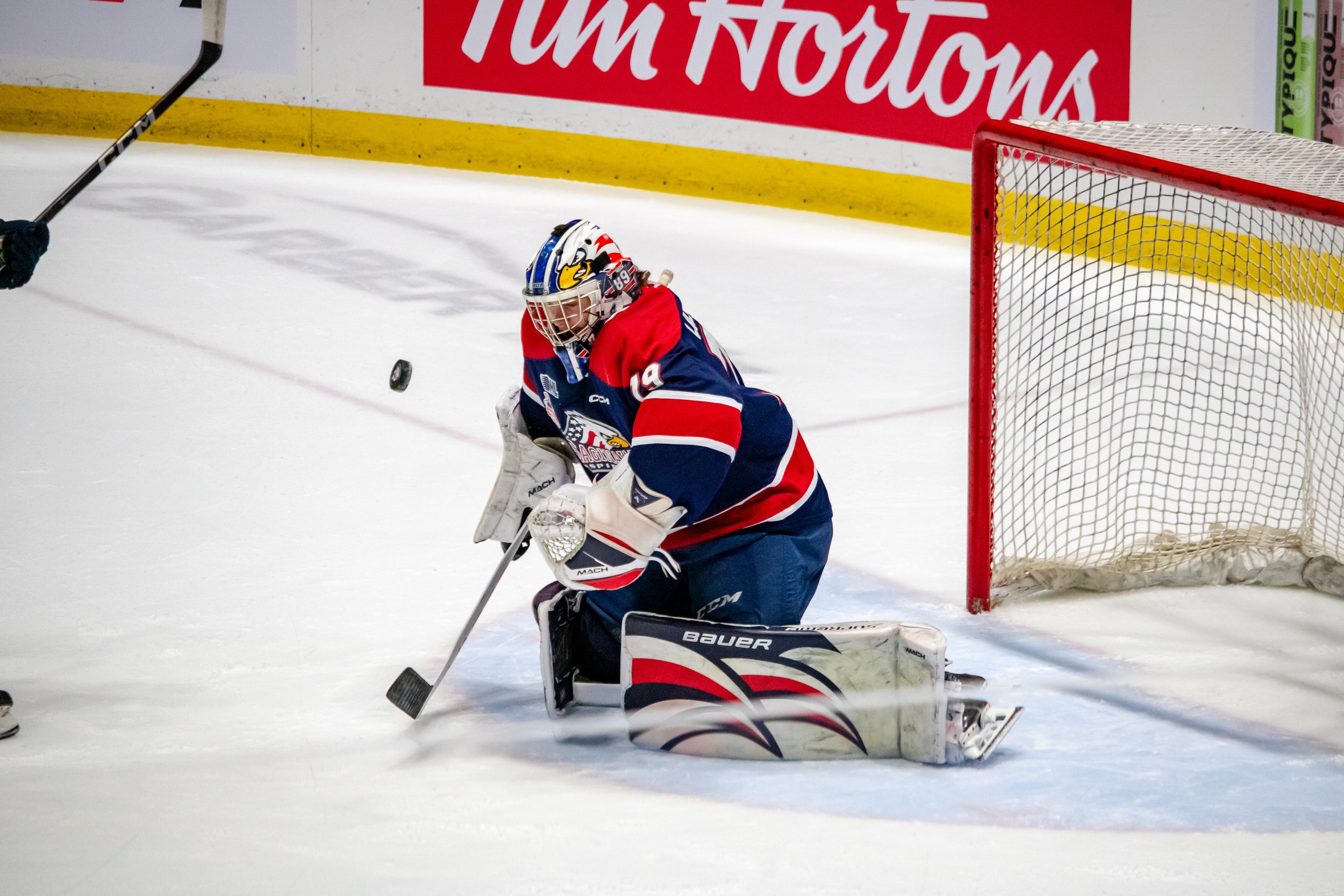Hockey goalie in protective gear, kneeling on the ice, catching a puck near the net during a game.