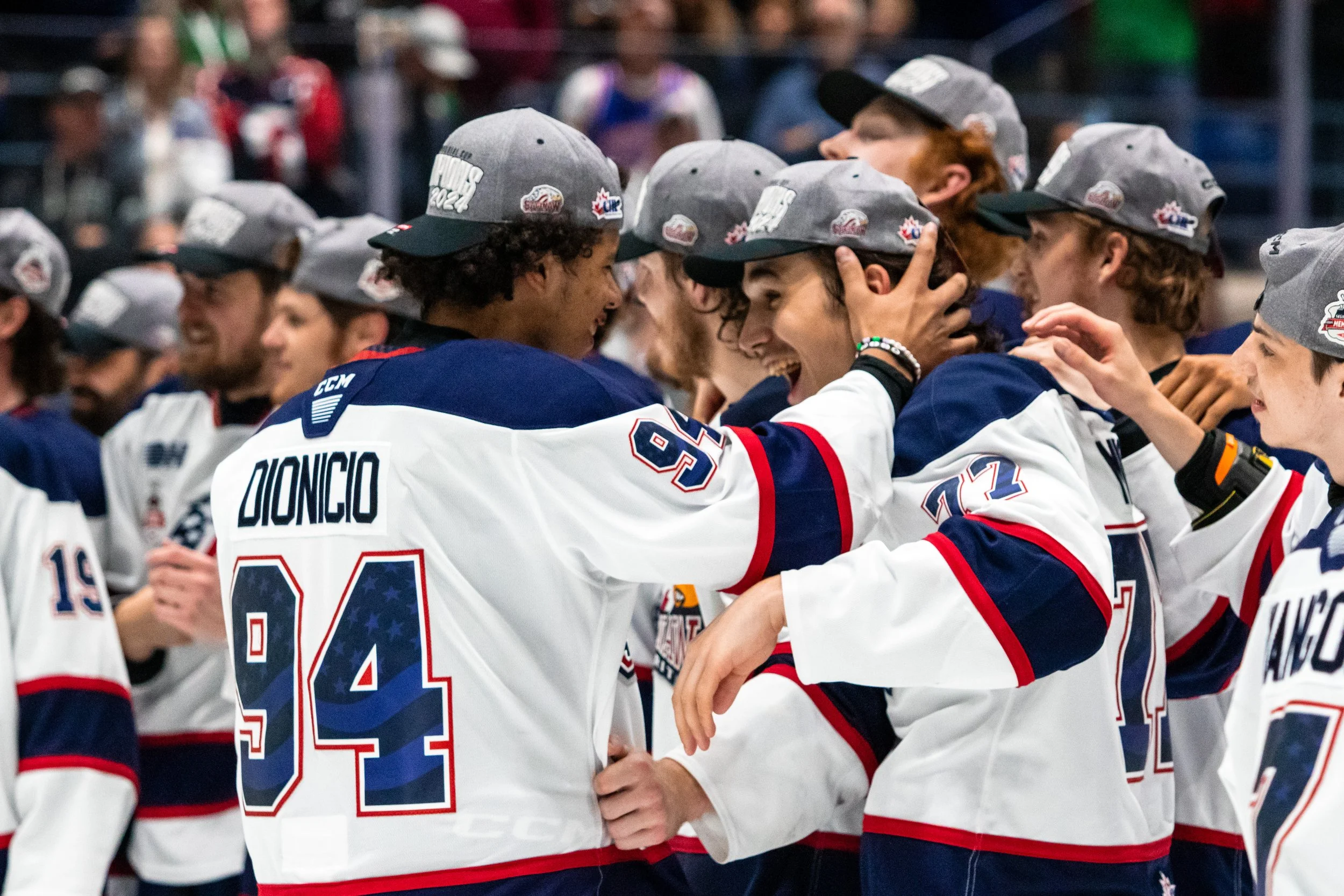 Ice hockey players in white jerseys with blue and red accents celebrating together during a game or event, with gray caps on their heads.