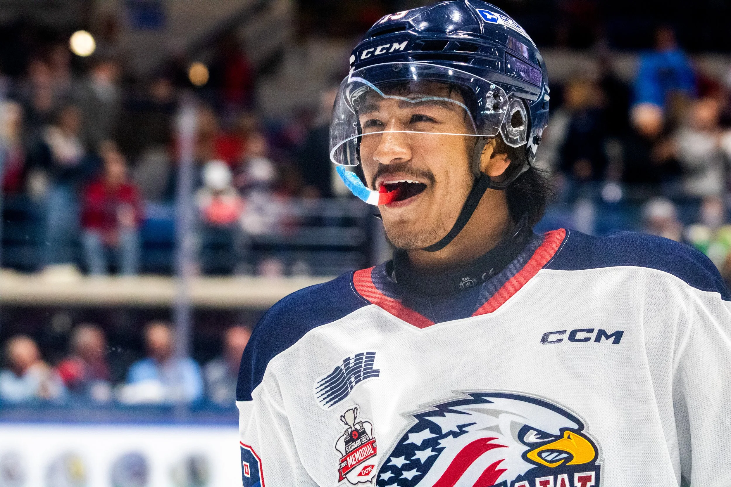 Hockey player wearing a white jersey with an eagle logo, navy blue helmet with a clear face shield, and a red whistle in his mouth, on an ice rink with spectators in the background.