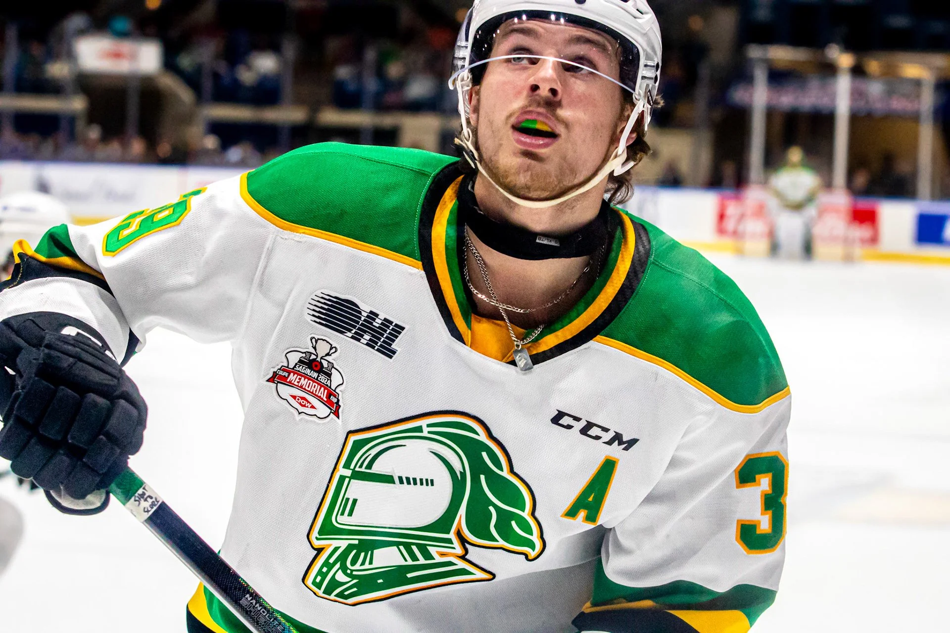 Hockey player in white and green jersey on ice rink, wearing helmet and gloves, with other players in the background.