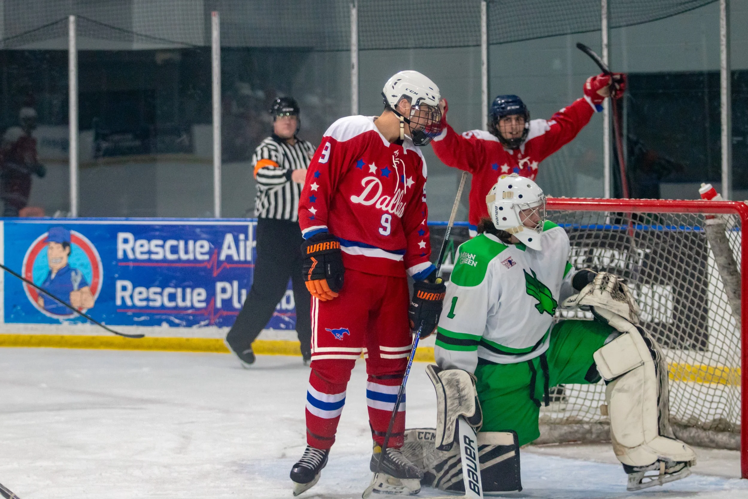 An ice hockey game showing a player in a red jersey, number 9, with his head bowed, standing after scoring or missing a shot. The goalie in green and white gear is on the ice, and a referee is in the background. Another player in a red jersey is cele