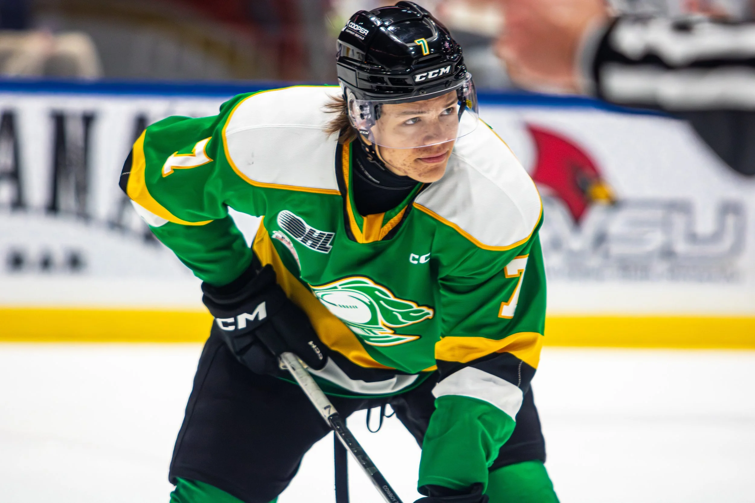 A hockey player in a green, white, and yellow jersey with the number 7, wearing a black helmet with a clear face shield, on the ice during a game.