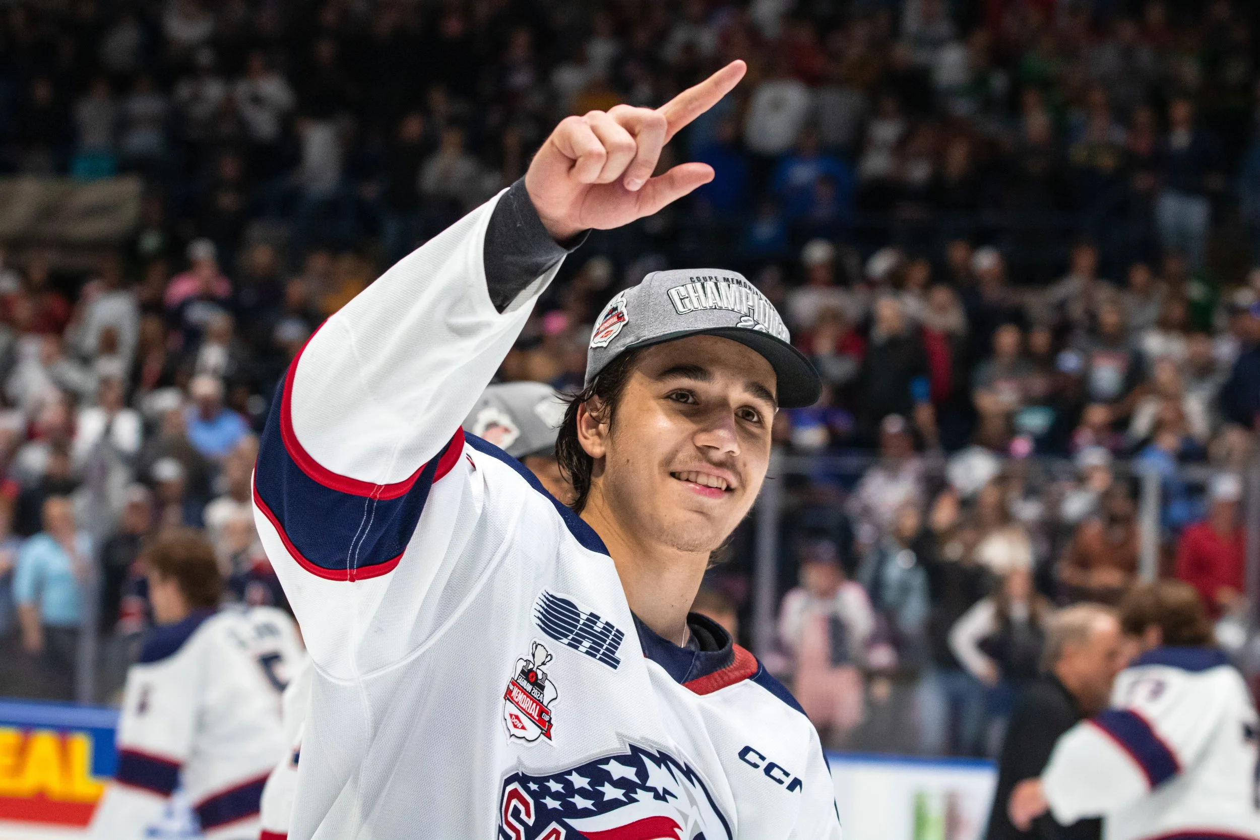A young male ice hockey player in a white, red, and blue jersey is smiling and raising his hand with his index finger pointing upward at a hockey rink during a game, with a large crowd of spectators in the background.