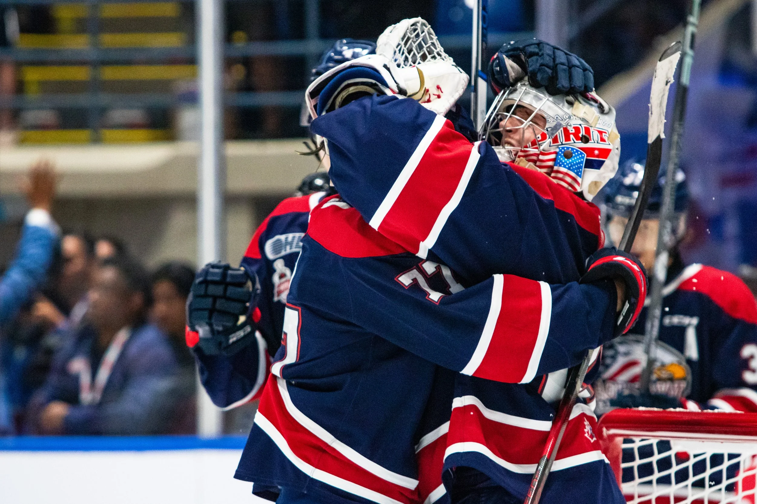 Two ice hockey players in red, white, and blue uniforms hug each other on the ice rink after a game, celebrating.