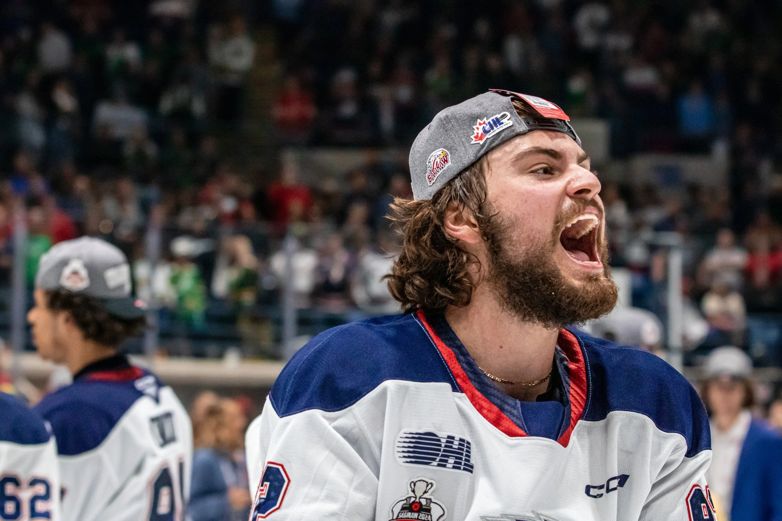 A hockey player with long brown hair and a beard, wearing a gray cap backwards and a hockey jersey, celebrating during a game or event with a cheering crowd in the background.