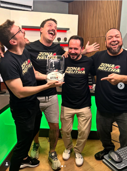 Quatro homens sorrindo, segurando um troféu, todos usando camisetas pretas com o logo 'Zona Neutra', em um ambiente com decoração moderna e iluminação colorida.