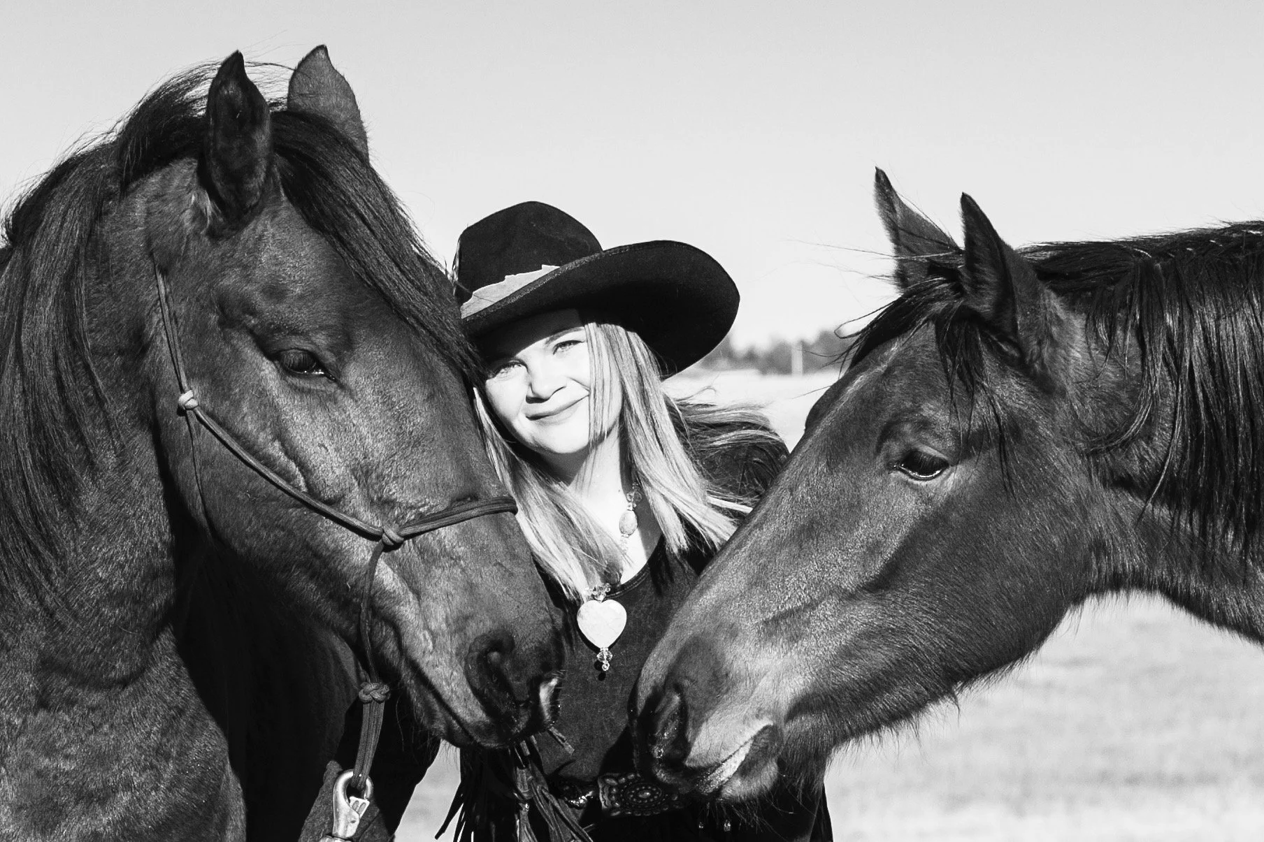 A girl with long hair wearing a cowboy hat standing between two horses, touching their faces in a field.