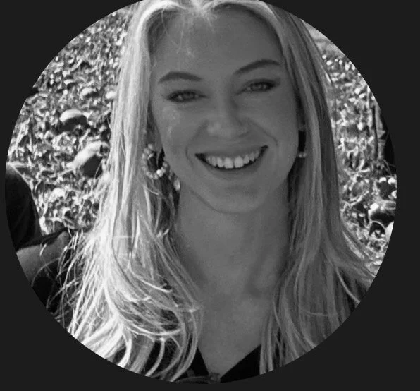 A smiling young woman with long blonde hair, wearing hoop earrings, outdoors with trees in the background.