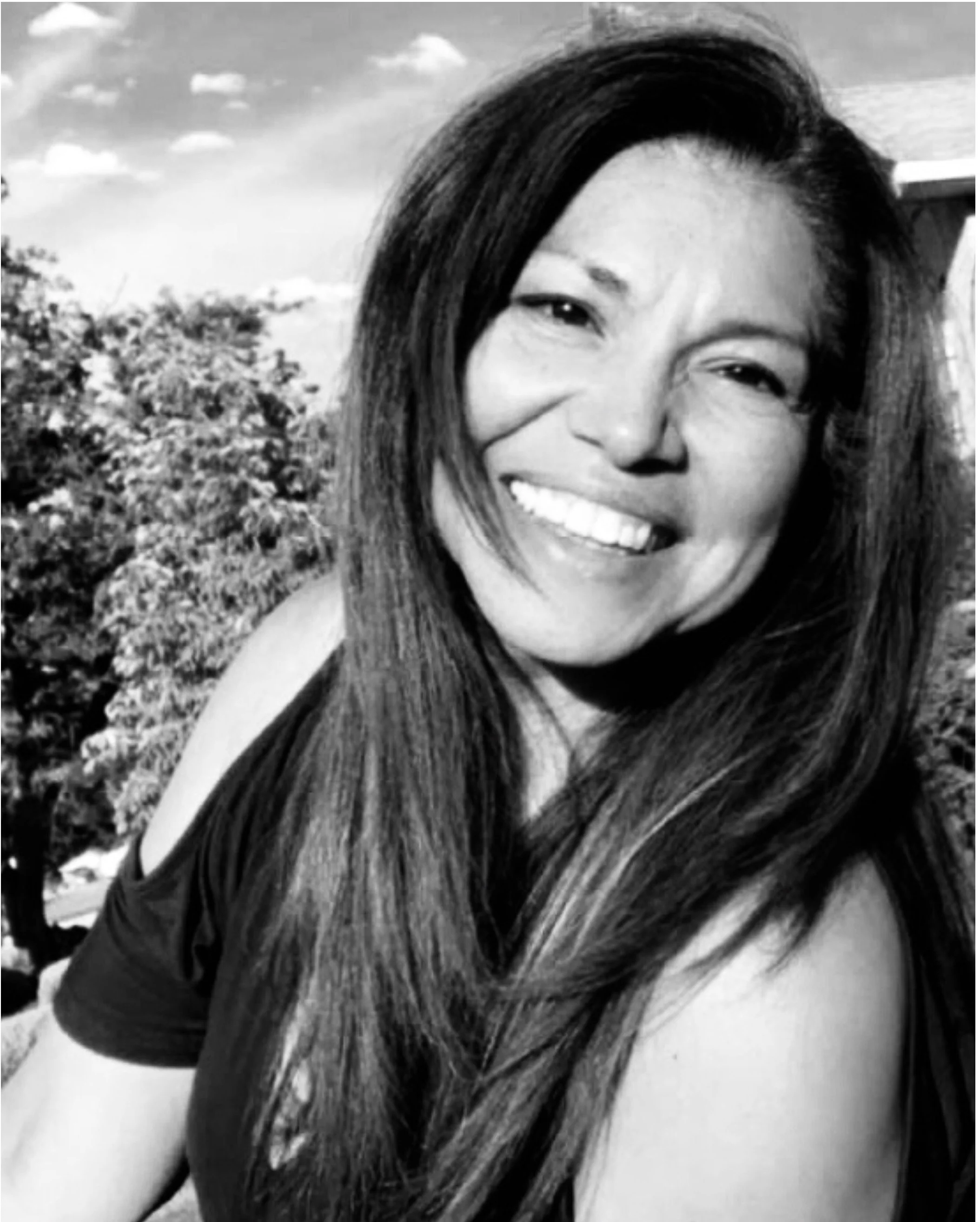 A woman smiling outdoors on a sunny day, with trees and clouds in the background.