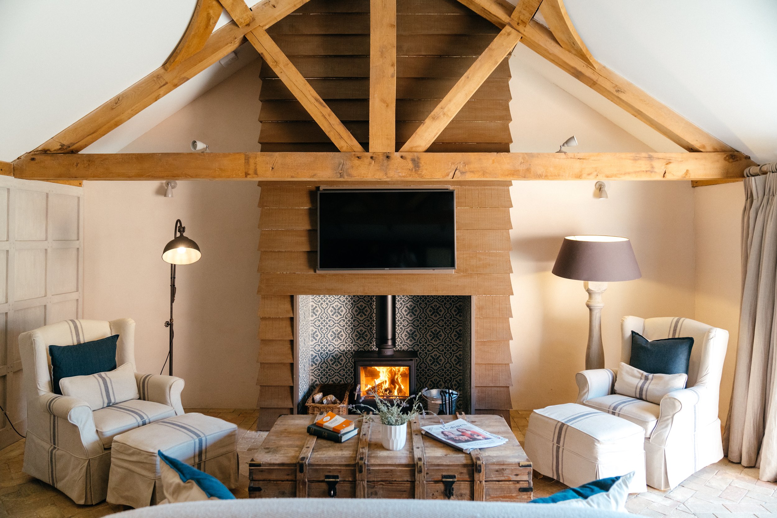Cozy living room with a central fireplace beneath a wall-mounted TV, surrounded by two white armchairs with blue pillows, a wooden coffee table with books and a potted plant, a tall floor lamp with a gray shade, and a floor lamp with a black shade. The room features exposed wooden ceiling beams and a wood-paneled accent. A window with light-colored curtains is visible on the right.