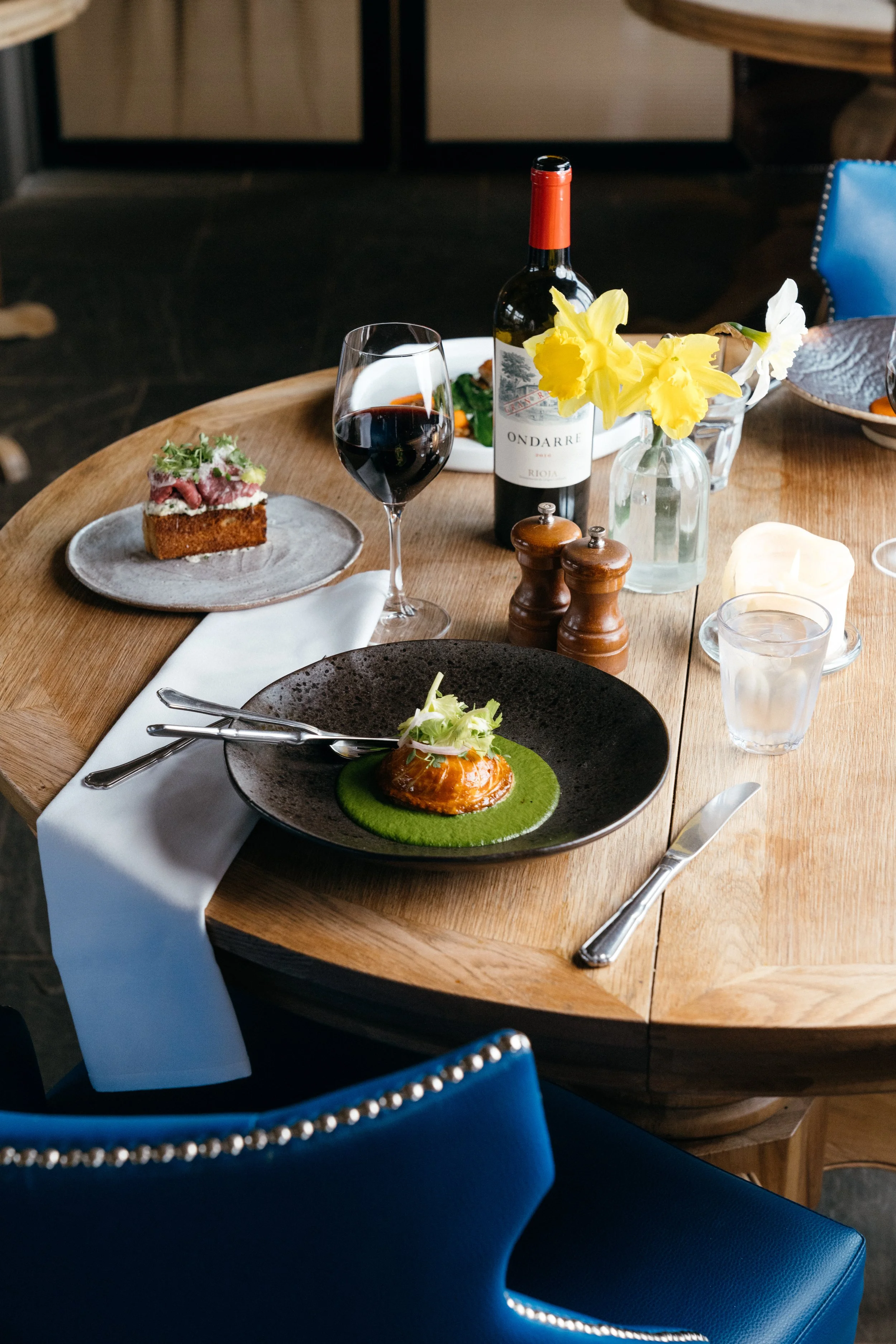 A wooden dining table set with a black plate featuring a gourmet dish, a glass of red wine, a bottle of wine, a small plate with a bread item, a glass of water, a candle, and a centerpiece with yellow and white flowers. Blue chairs surround the table.