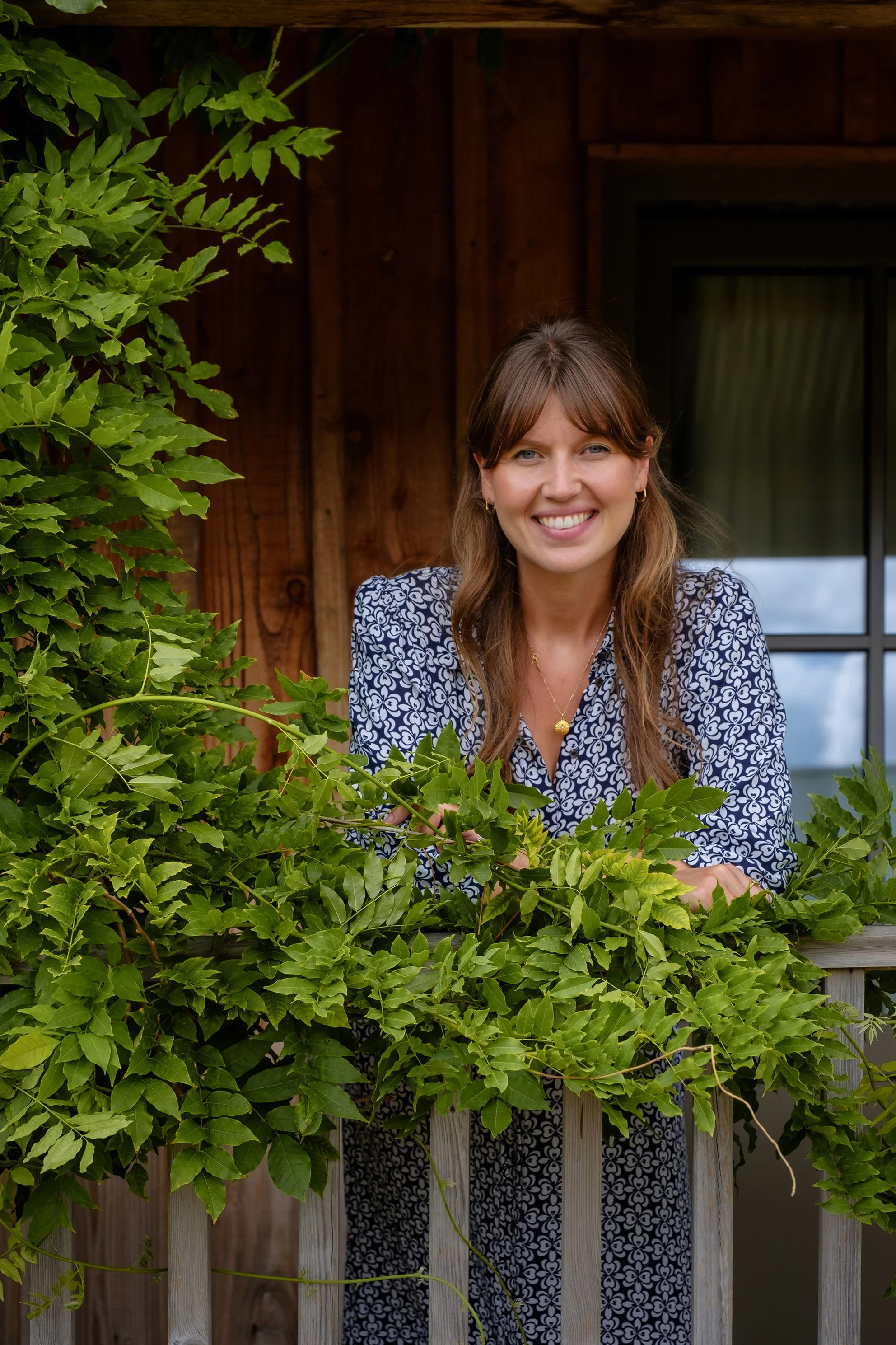Steph Jones, Founder of WanderFolk. A woman with brown hair in loose waves, wearing a patterned blue and white blouse and gold jewellery, smiles while leaning on a wooden railing amidst greenery in front of a rustic wooden building