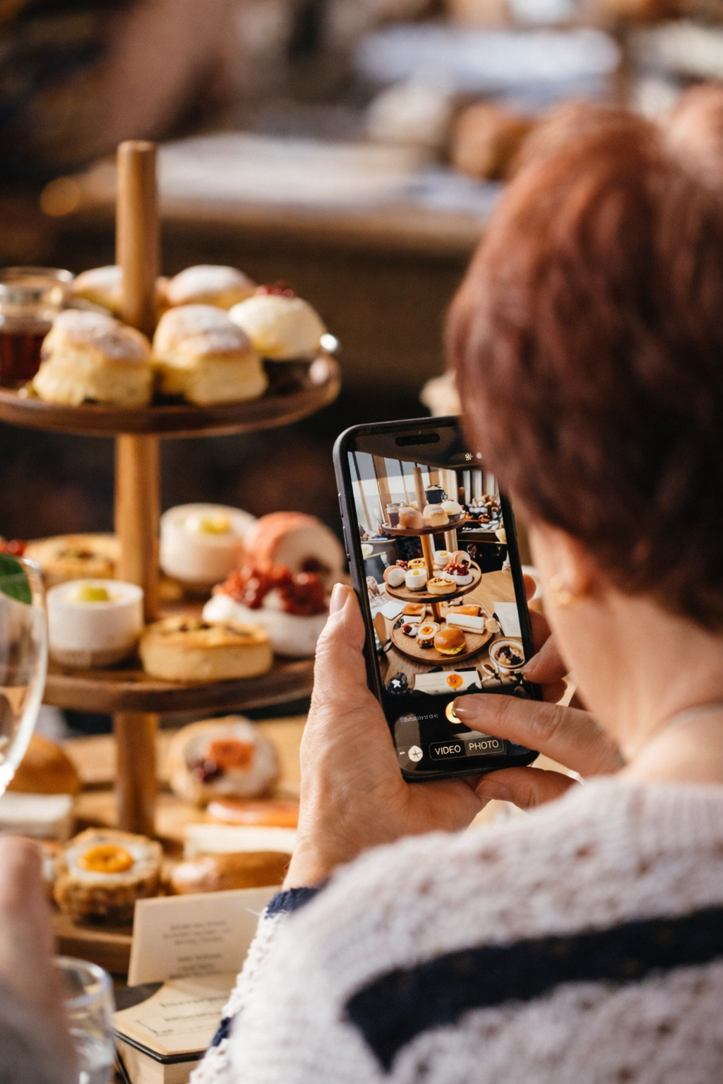 Person taking a photo of a dessert display with a smartphone. The display includes cookies, scones, and pastries on a tiered wooden stand.