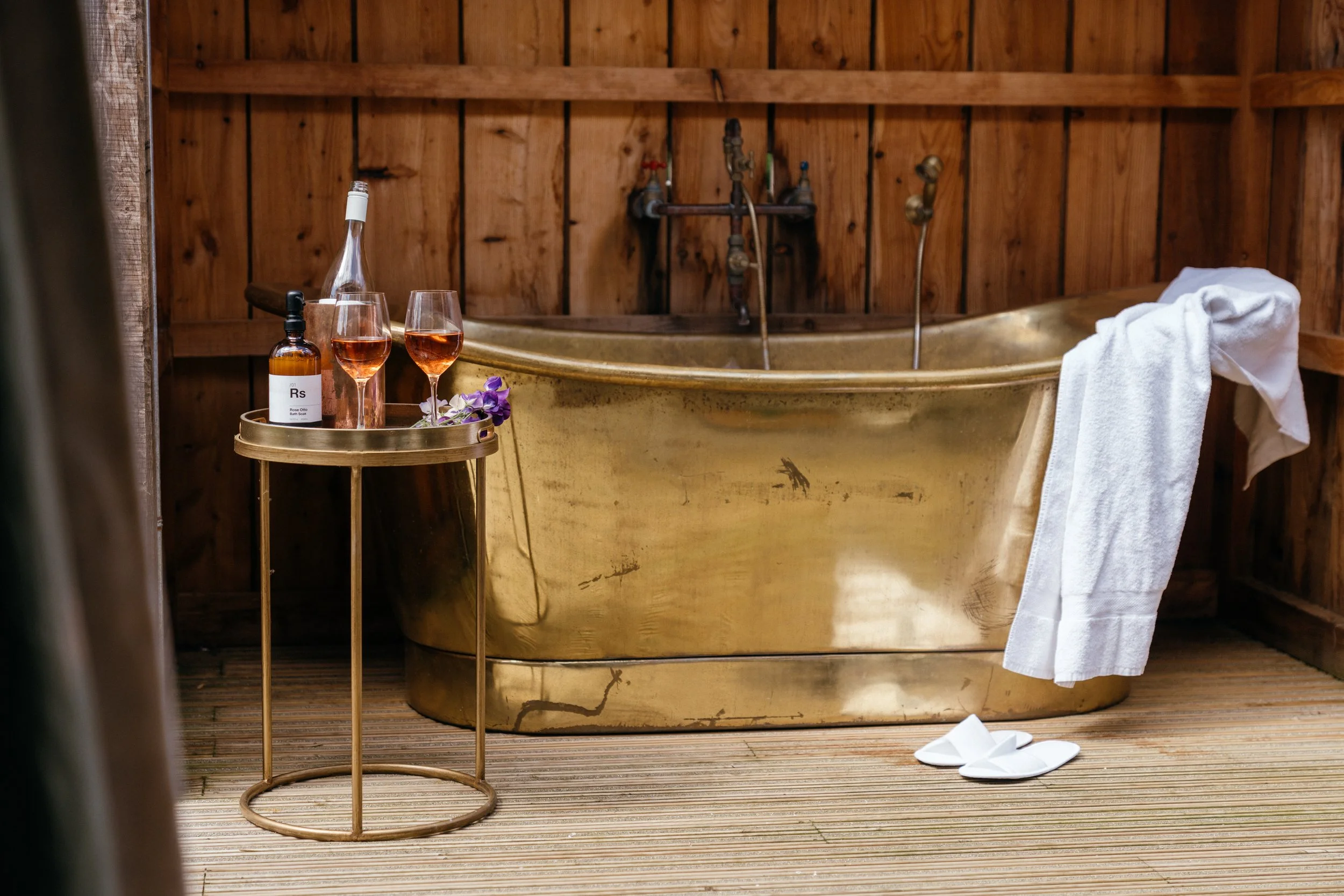 A bronze bathtub in a wooden shed or bathroom with a white towel hanging over the edge, a pair of white slippers on the wooden floor, and a small round table with two glasses of rosé wine, a bottle of wine, a dark bottle of soap or lotion, and some purple flowers.