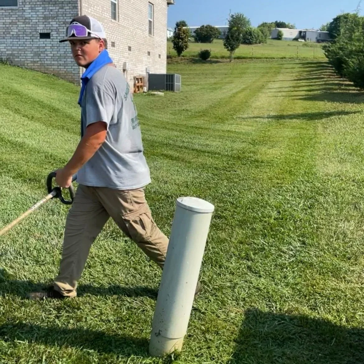 Man wearing gray shirt, khaki pants, cap, sunglasses, and a blue bandana around his neck, standing on a grassy yard holding a weed trimmer near a white pole, with a house and trees in the background.