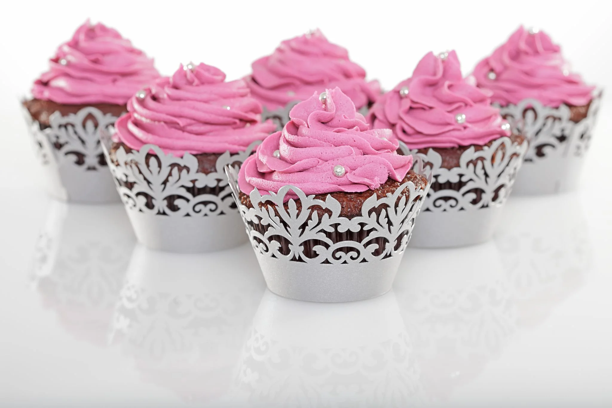 Five cupcakes with pink frosting in ornate silver wrappers on a reflective white surface.