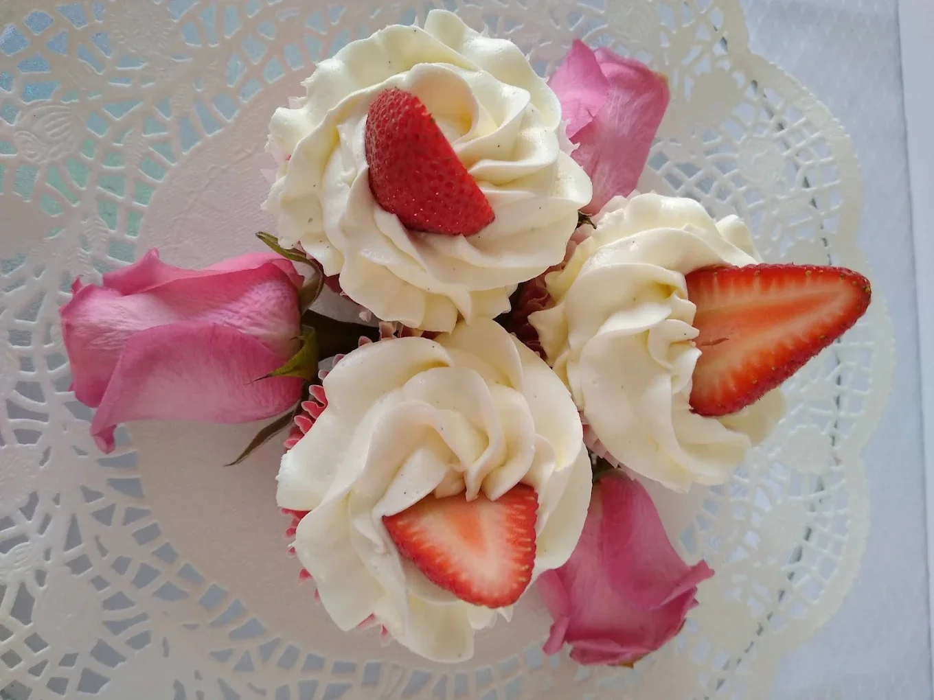 Cake decorated with white frosting, sliced strawberries, and pink rose petals, on a decorative lace-like doily.