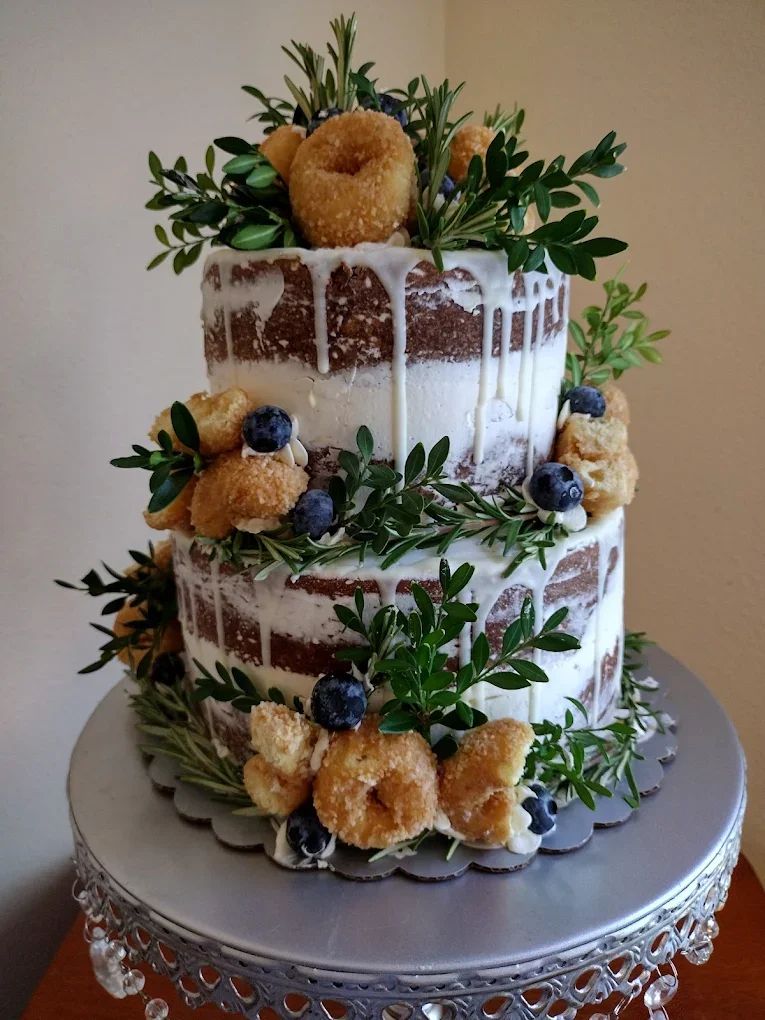 Two-layer naked cake decorated with blueberries, greenery, and mini doughnuts, with white icing drizzling down the sides, on a silver cake stand.