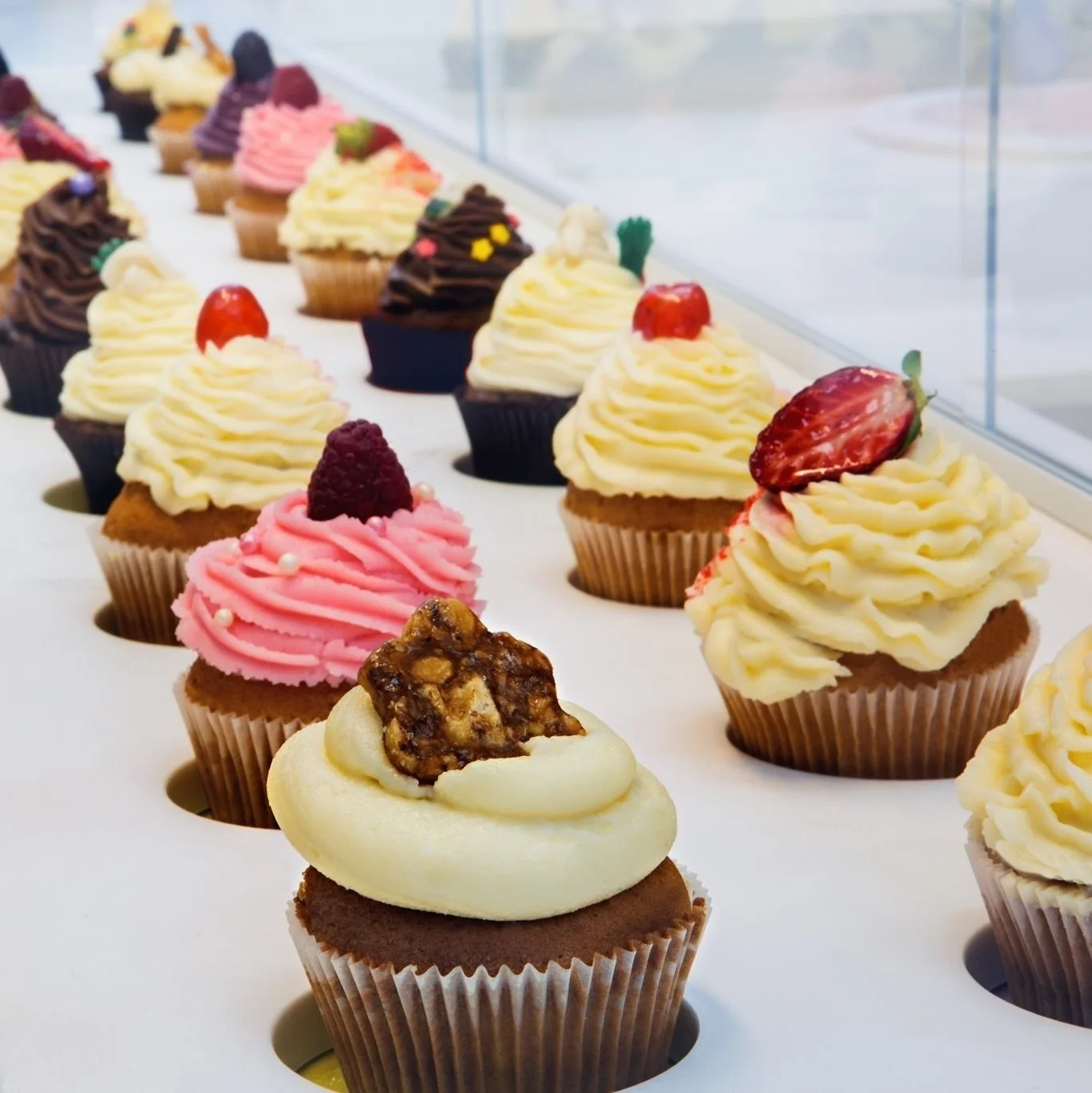 Display of various decorated cupcakes with different frosting styles and toppings such as strawberries, berries, and nuts.