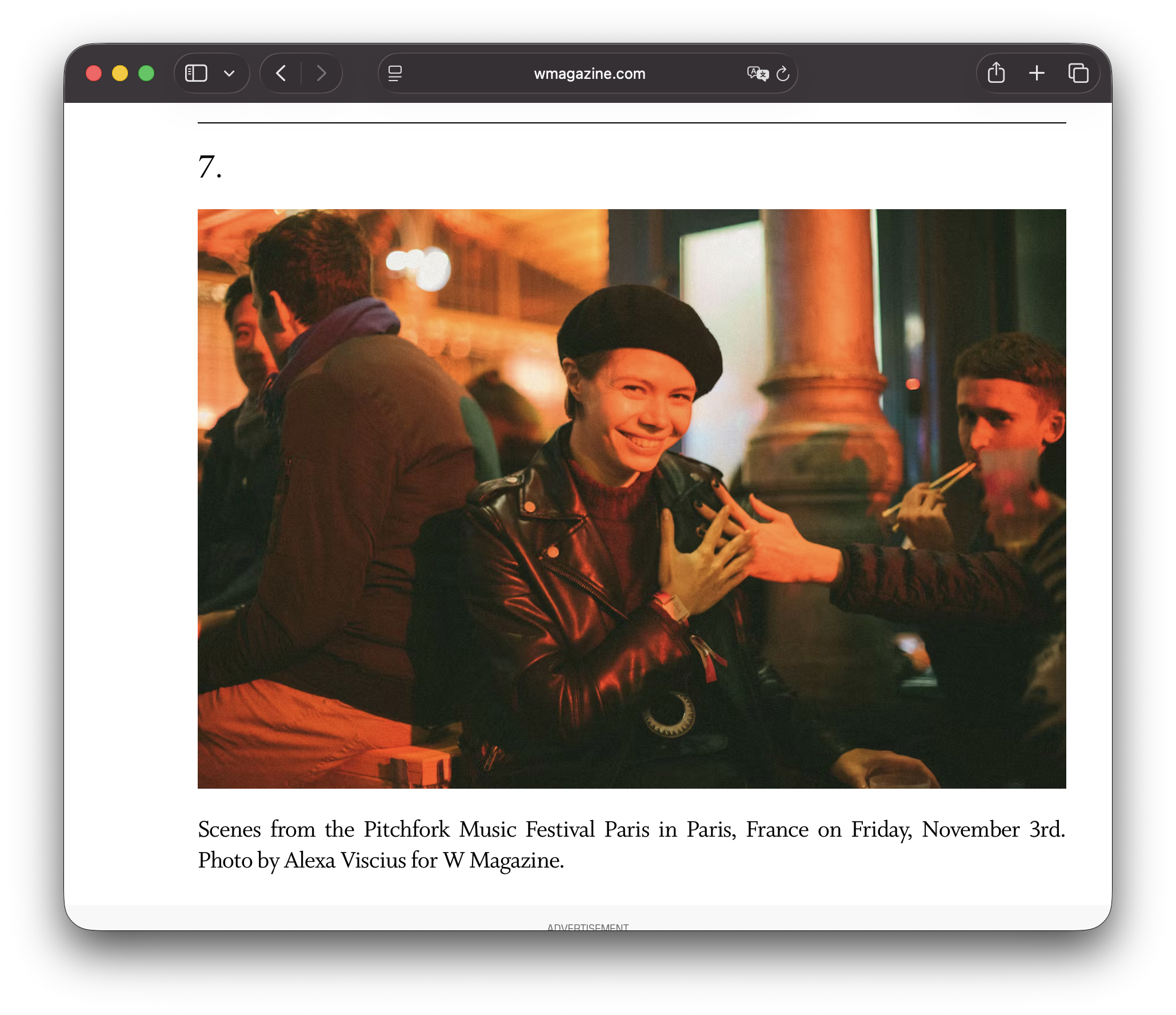 A group of people at the Pitchfork Music Festival in Paris, France, with a woman in the center wearing a black beret and leather jacket smiling as someone offers her a hand. The background is warm with orange and red lighting, and other attendees are visible.